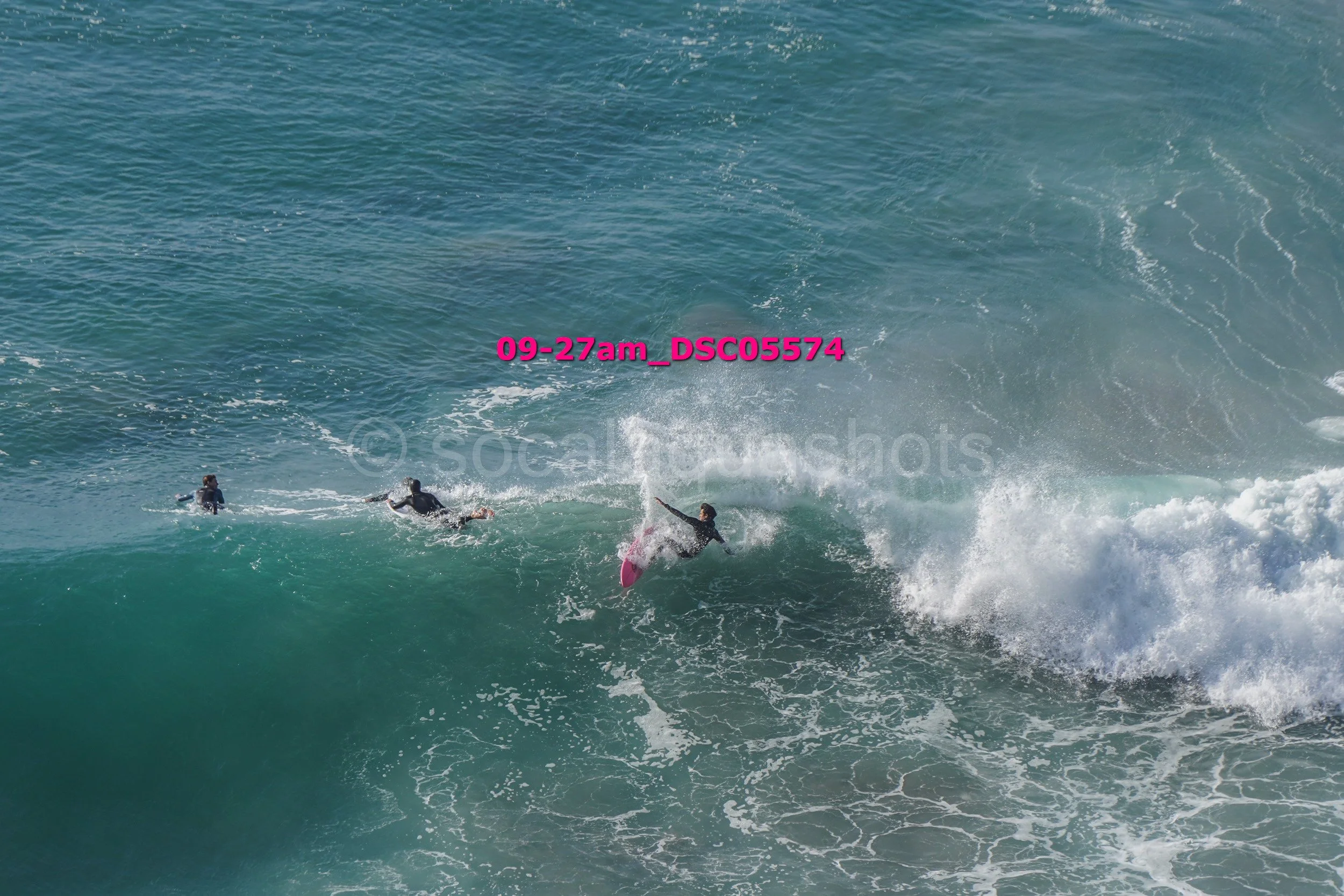 Three surfers riding a large wave in the ocean, one surfer falling off the board amid white water, while two others paddle nearby.