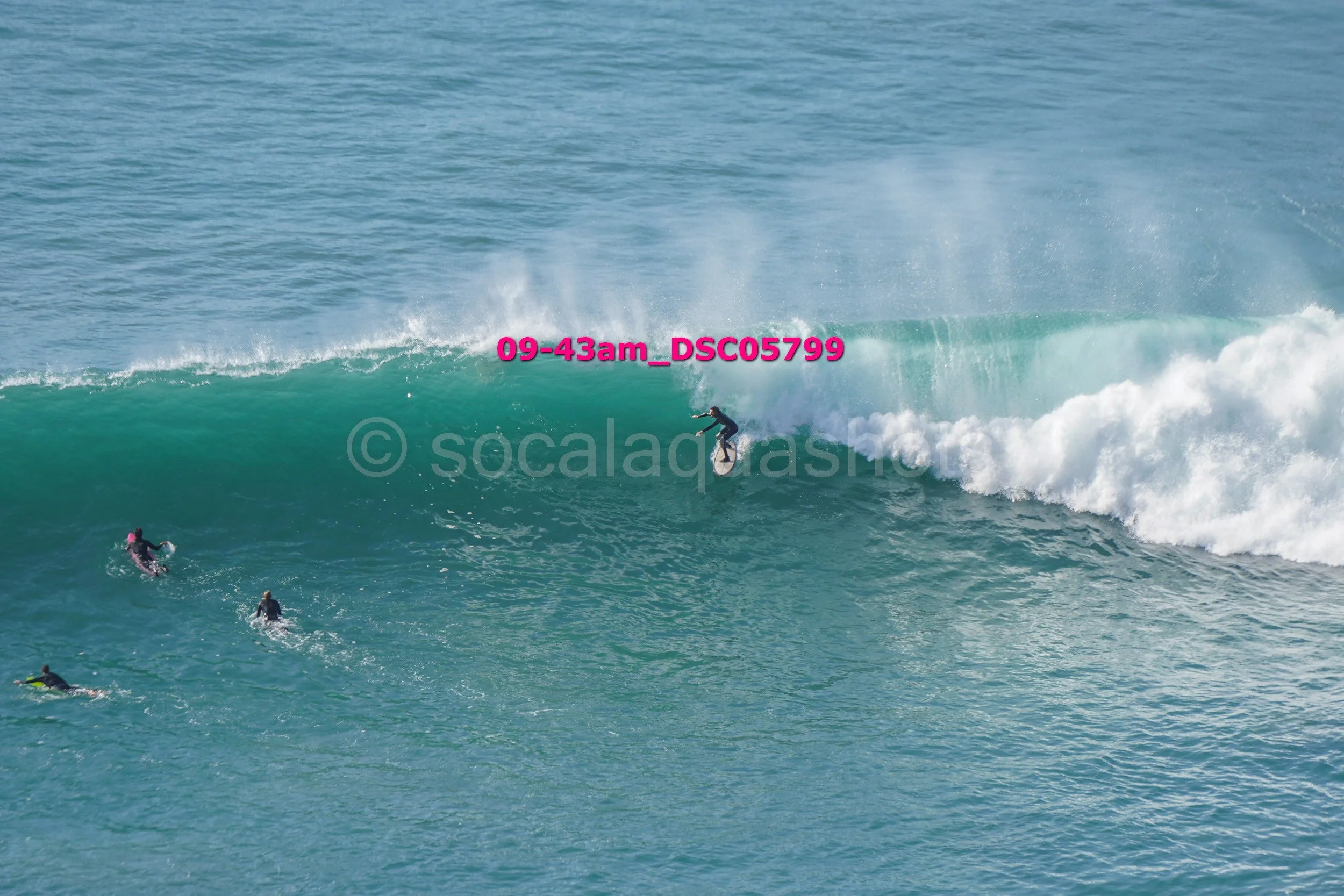 A person surfing on a large ocean wave with several other swimmers in the water nearby.