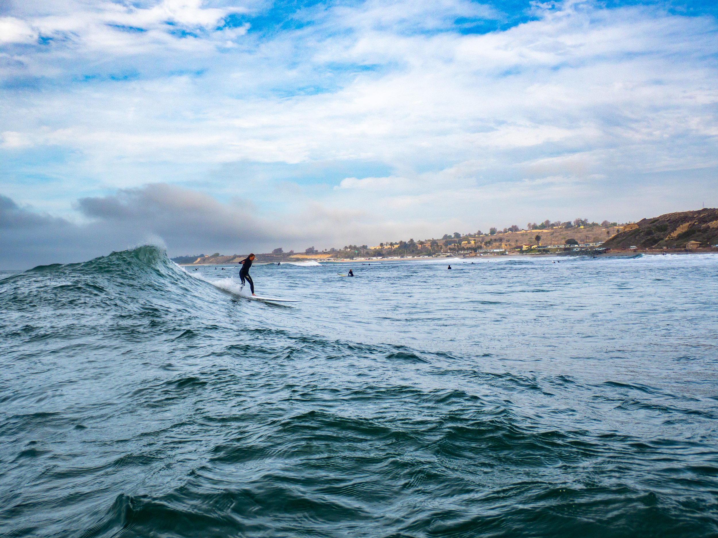 A person surfing on a wave in the ocean with other surfers in the distance and a coastal landscape in the background.