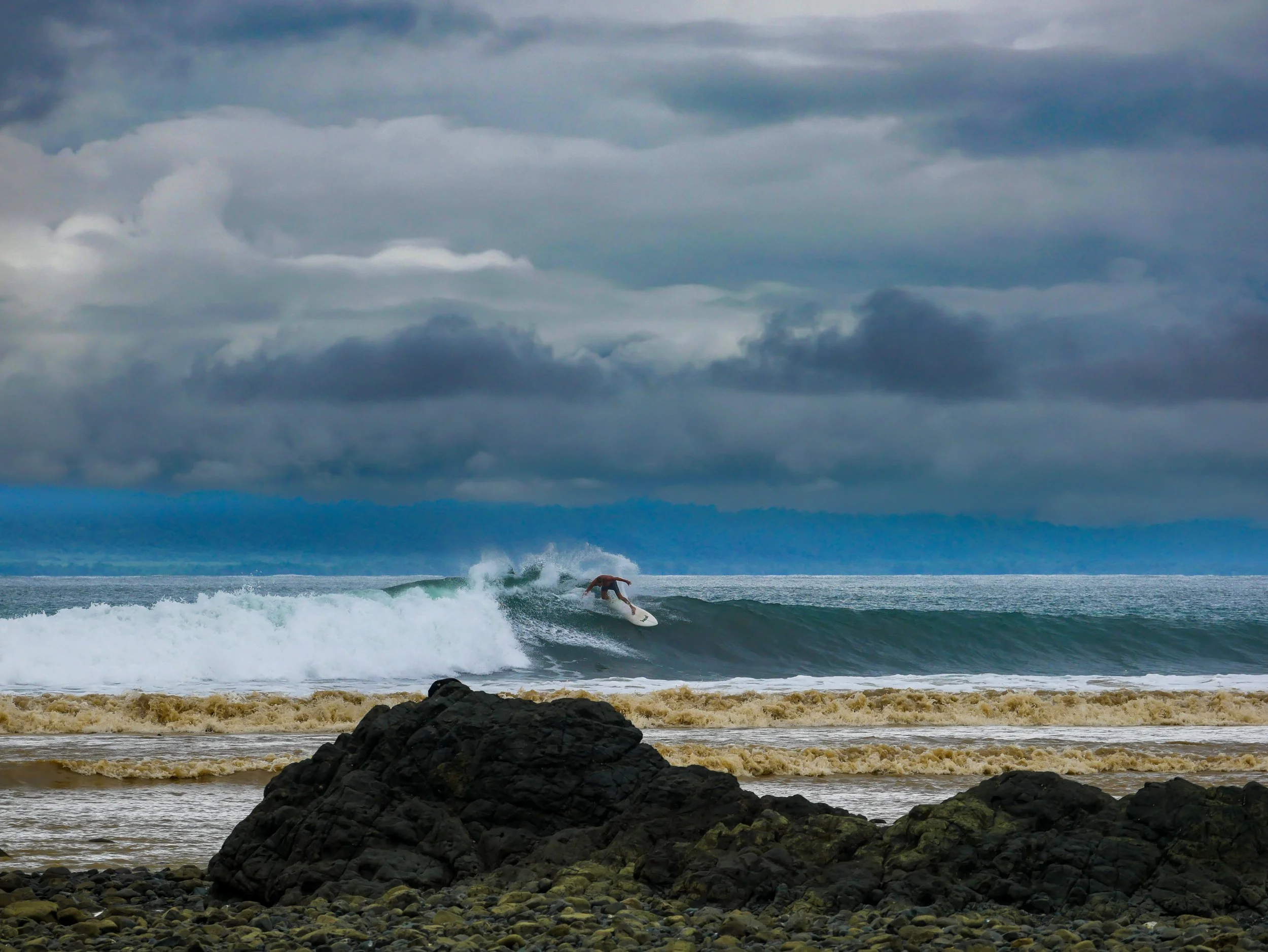 Surfer riding a wave near rocky shore, stormy sky overhead