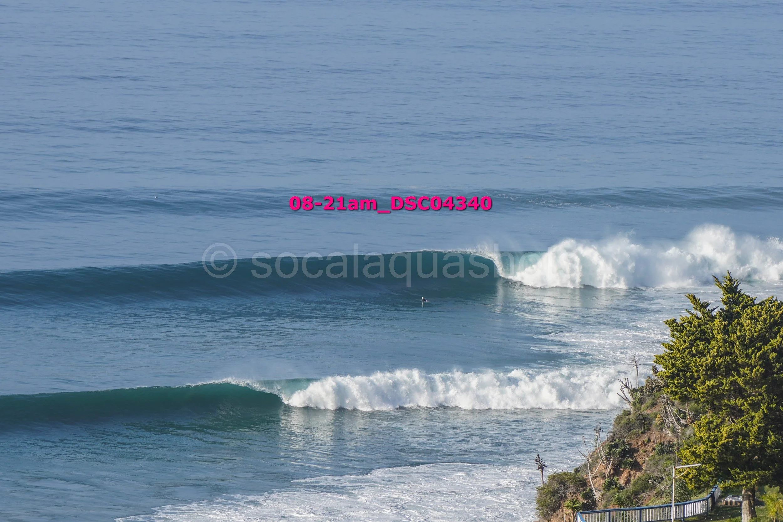 Ocean waves crashing near a rocky shoreline with trees, viewed from the coast on a clear day.