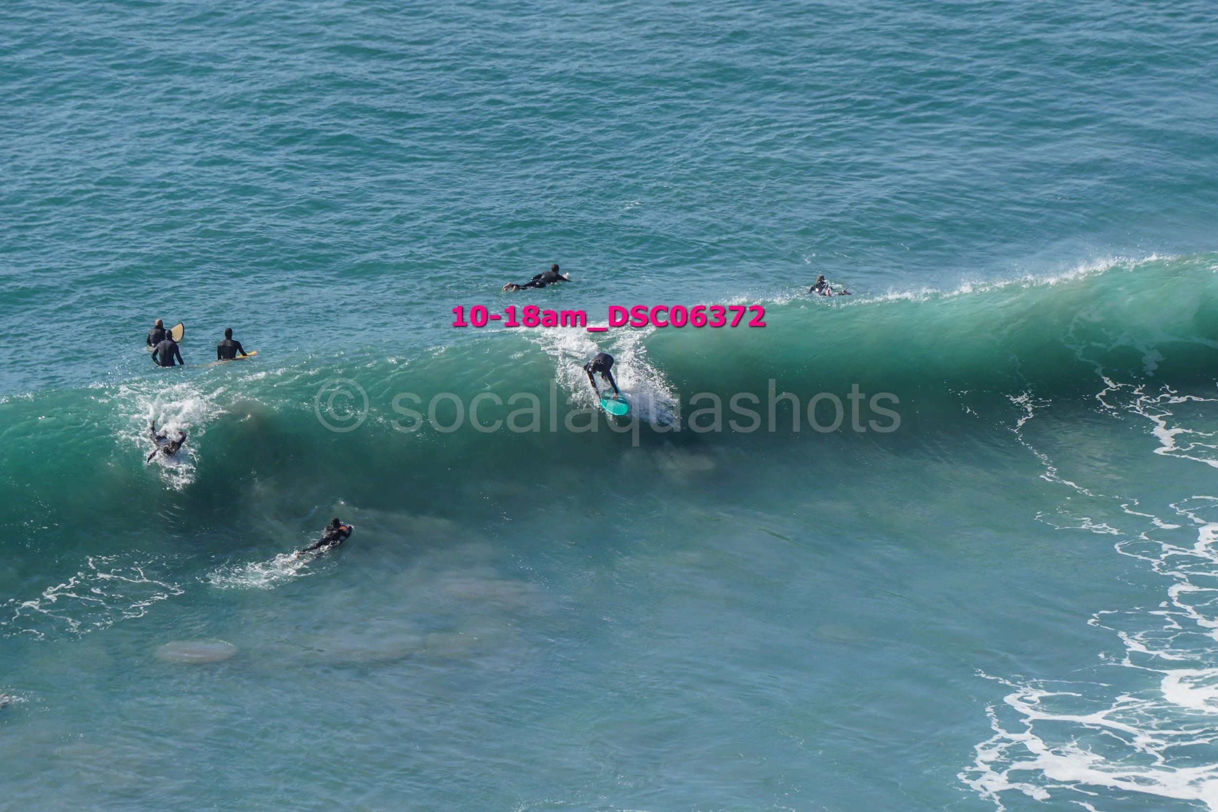 Surfers riding a large wave with others waiting in the water, wearing wetsuits in an ocean.