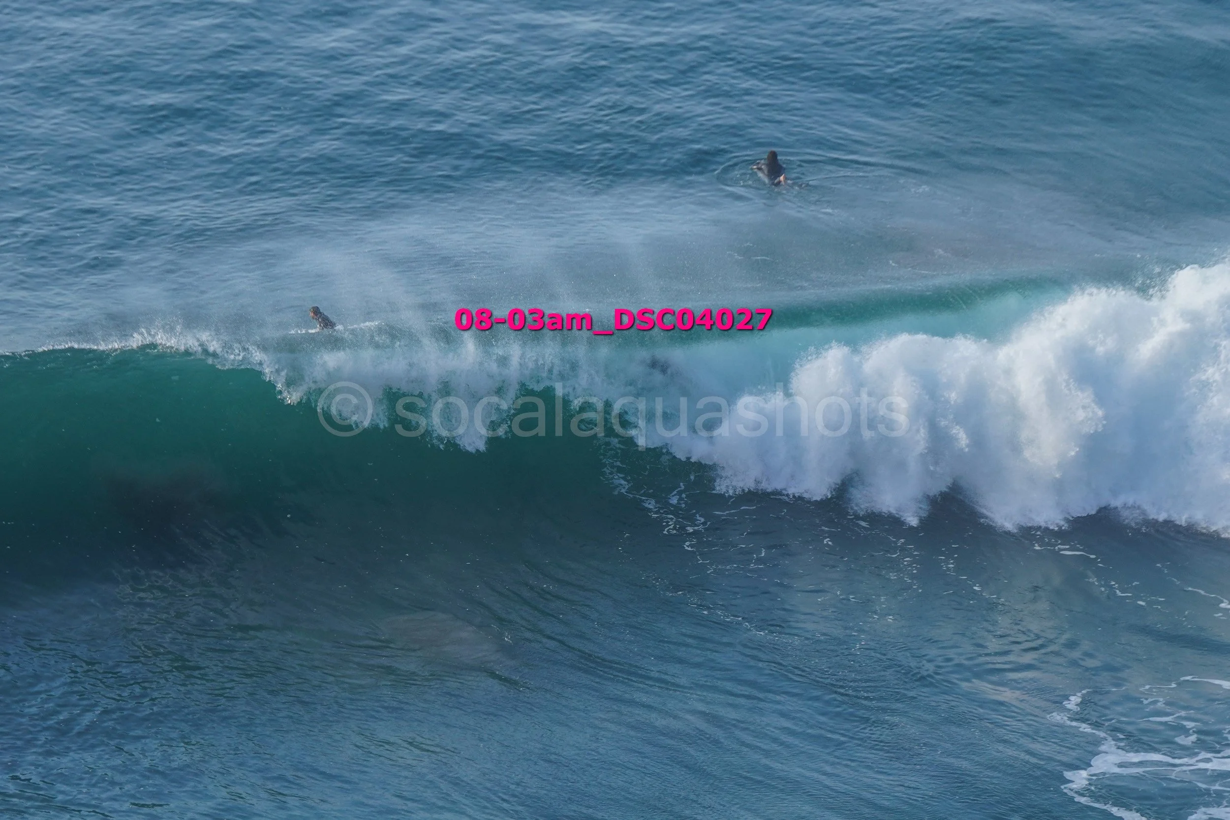 Two surfers are in the water with large waves, one partially visible behind the wave and the other farther back in the ocean.