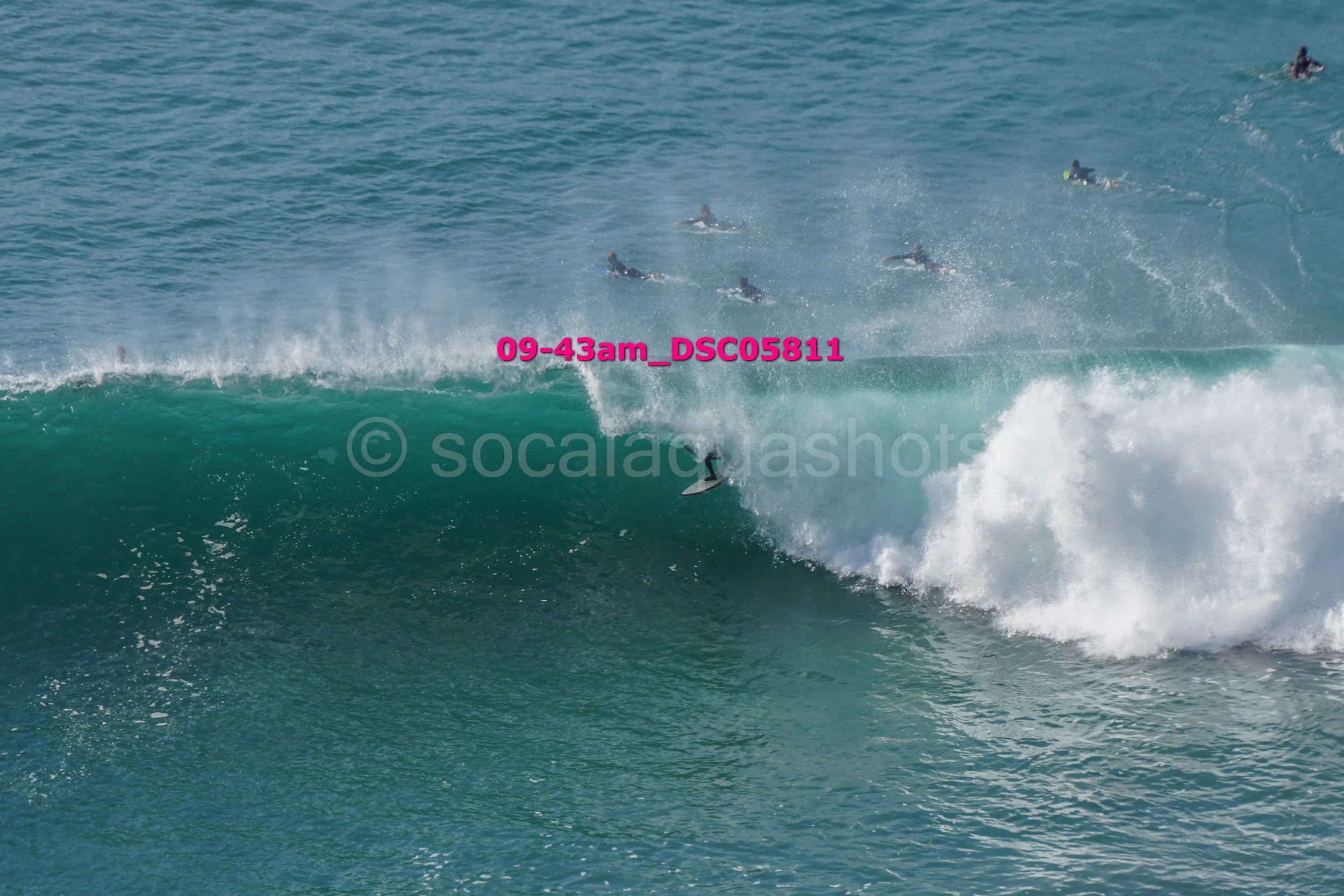 Surfer riding a large wave with several surfers swimming in the background.