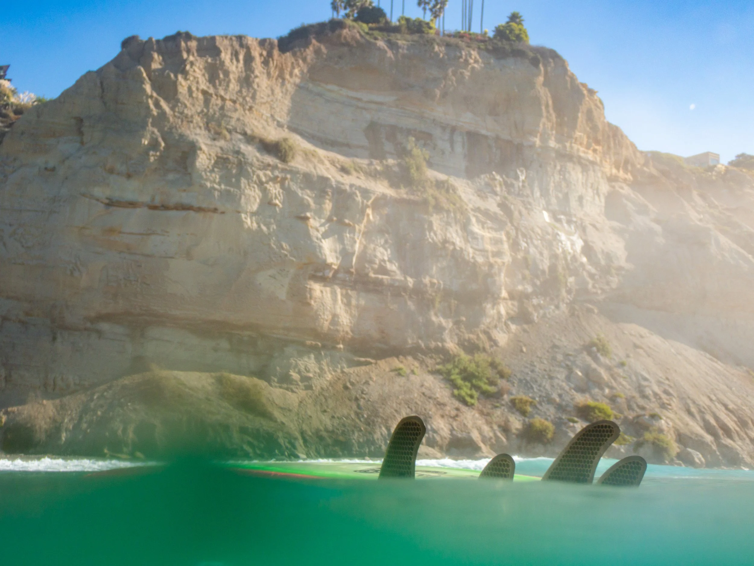 Footage of a surfboard with three fins in green water, with rocky cliffs and a blue sky in the background.