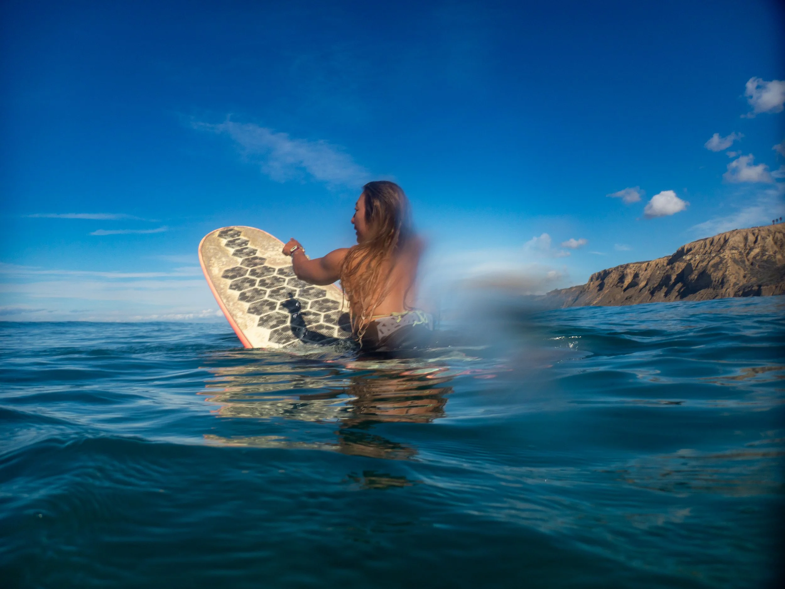 A woman in the ocean, holding a surfboard, with a rocky coastline and blue sky in the background.