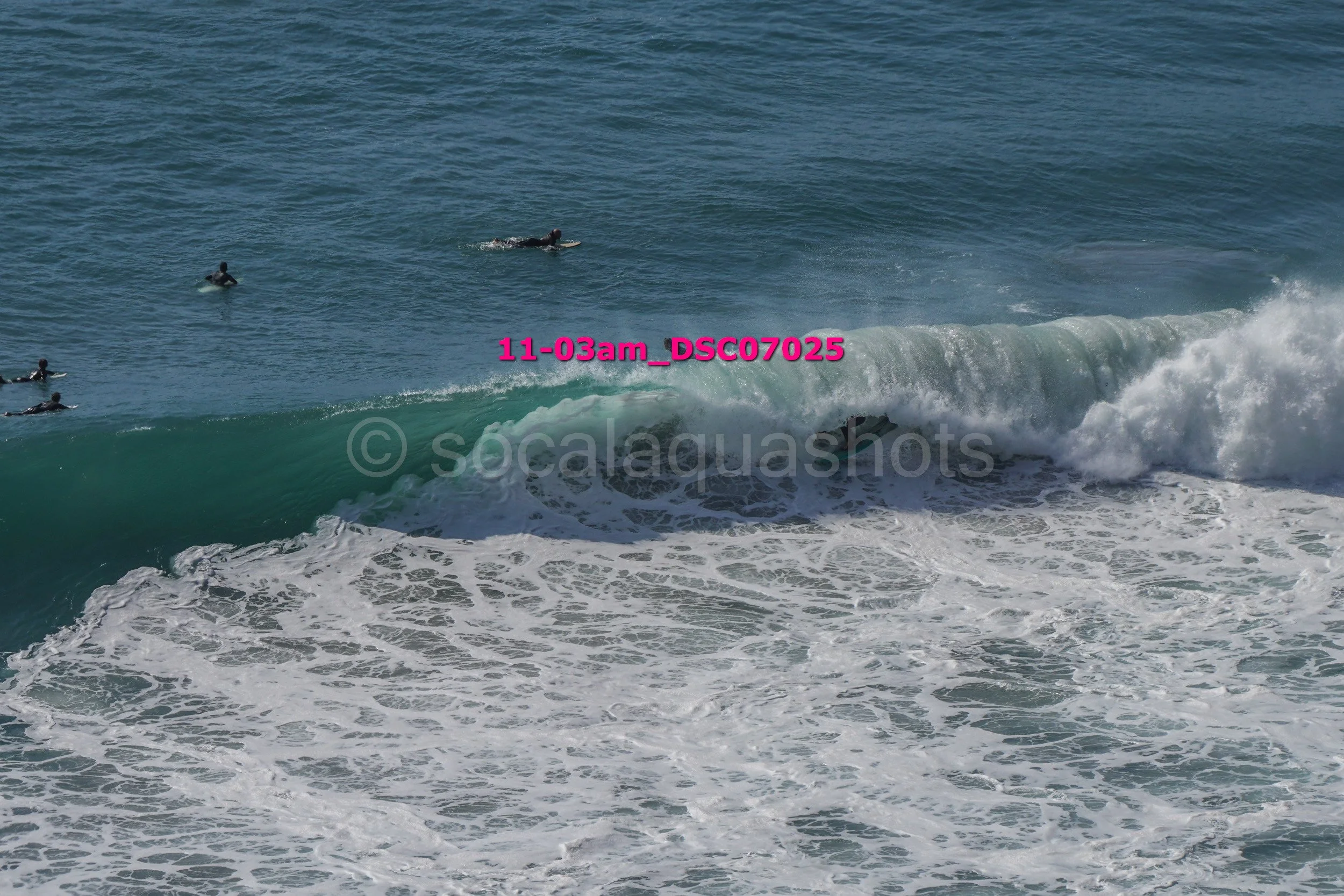 Several surfers in wetsuits riding and waiting for waves in the ocean near a breaking wave.