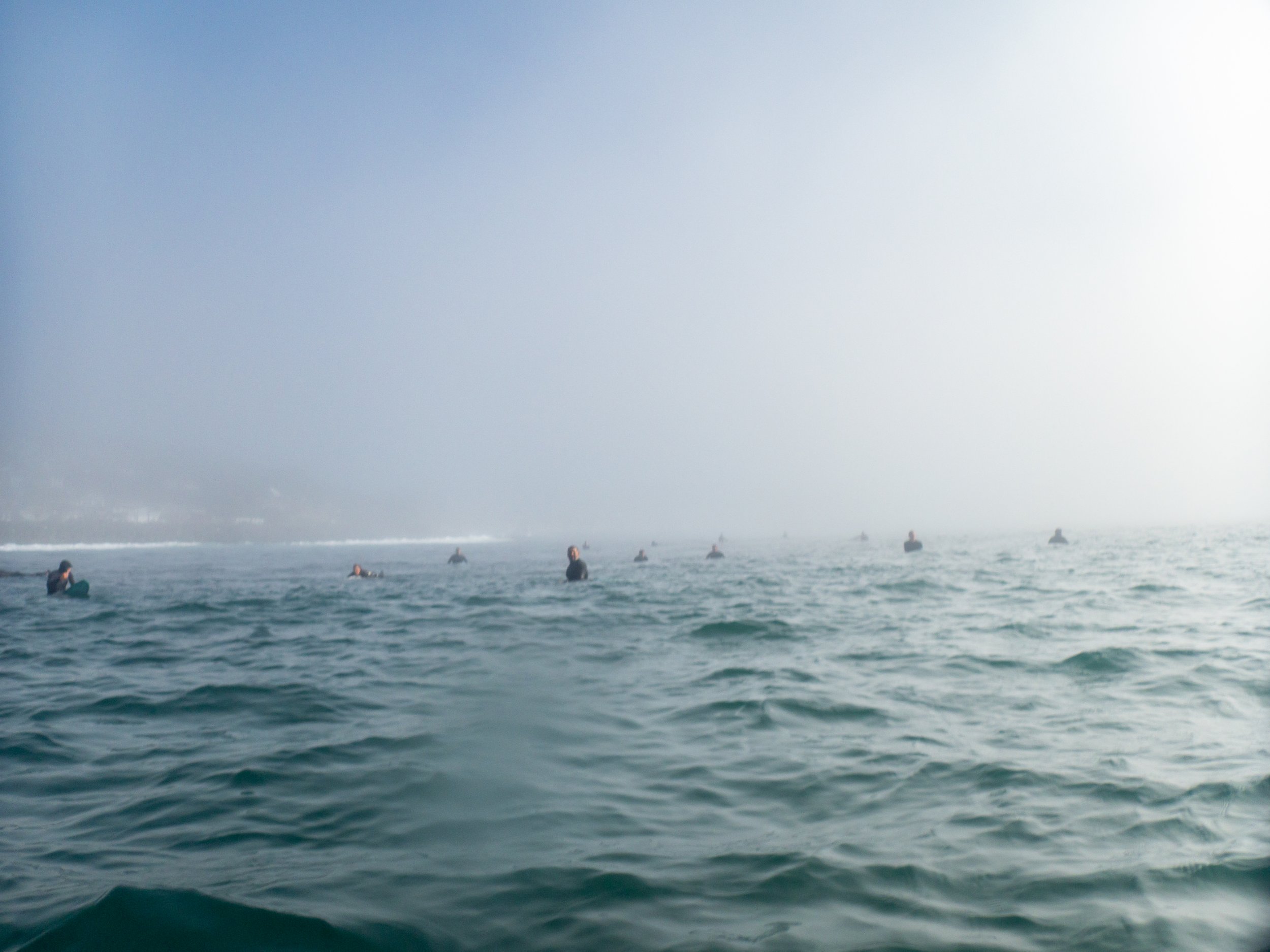 People in wetsuits sitting on surfboards in the ocean, with a foggy sky in the background.