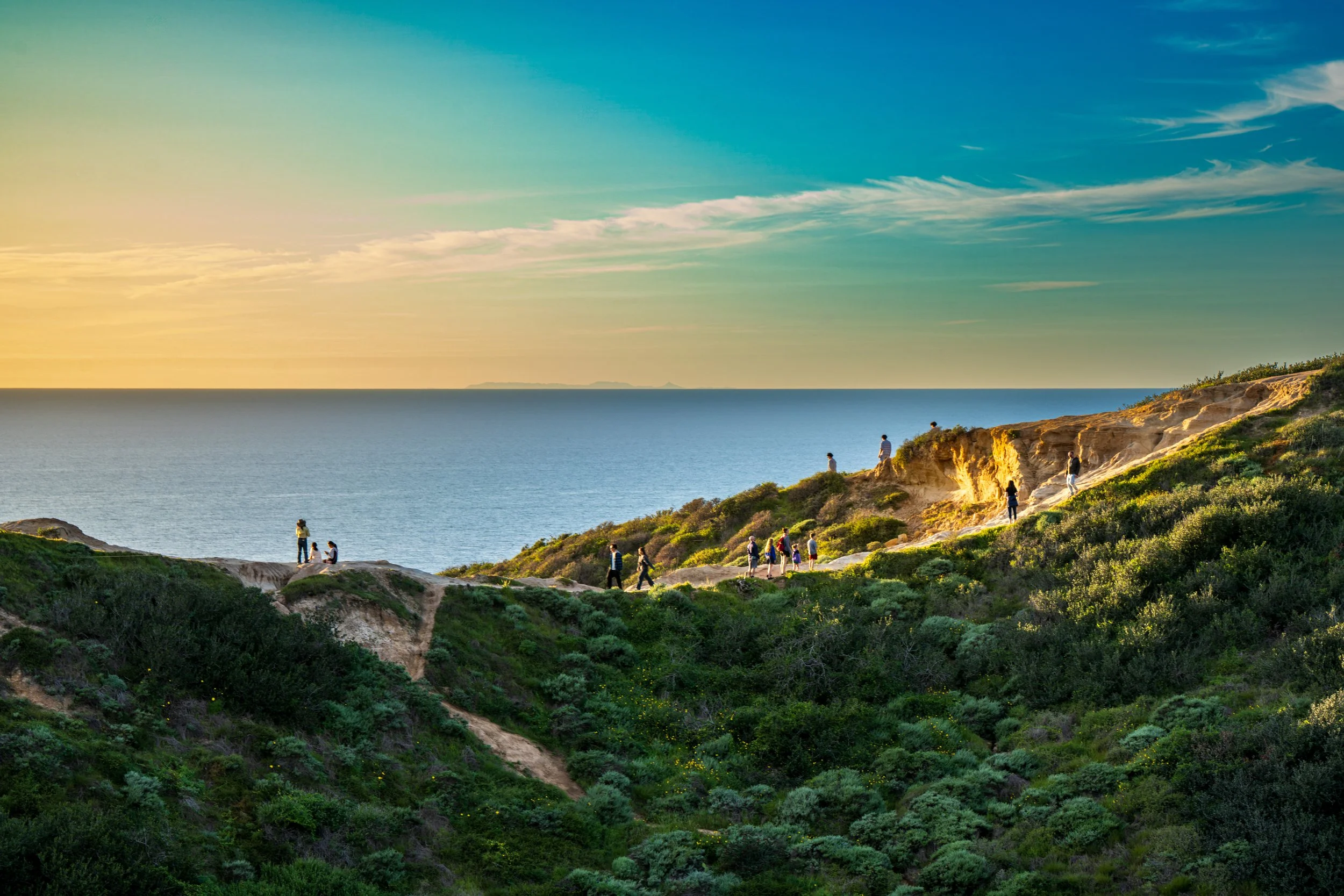 People walking and standing along a rugged, green hill overlooking the ocean at sunset.