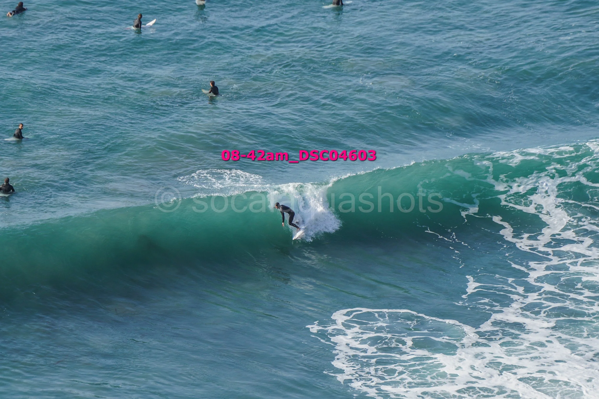 Surfer riding a wave in the ocean with several people in wetsuits and surfboards in the water nearby.