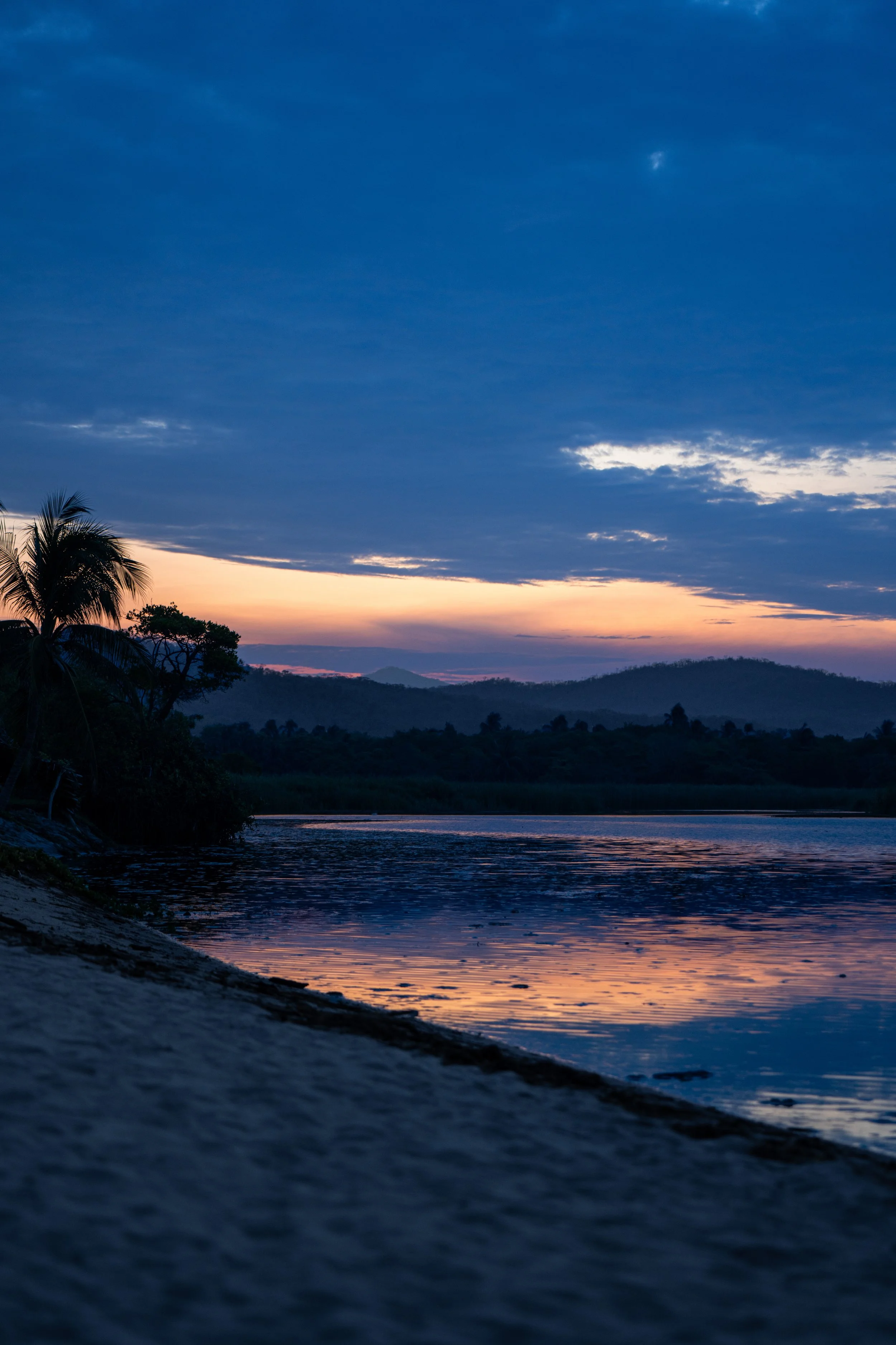 A scenic view of a river at dusk with a colorful sunset sky, silhouetted trees, and distant mountains.