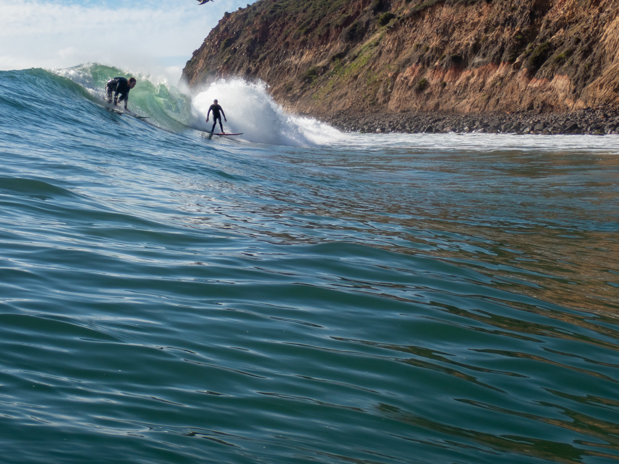 Two surfers riding a wave near a rocky cliffside, with one surfer in front and the other behind, in the ocean.