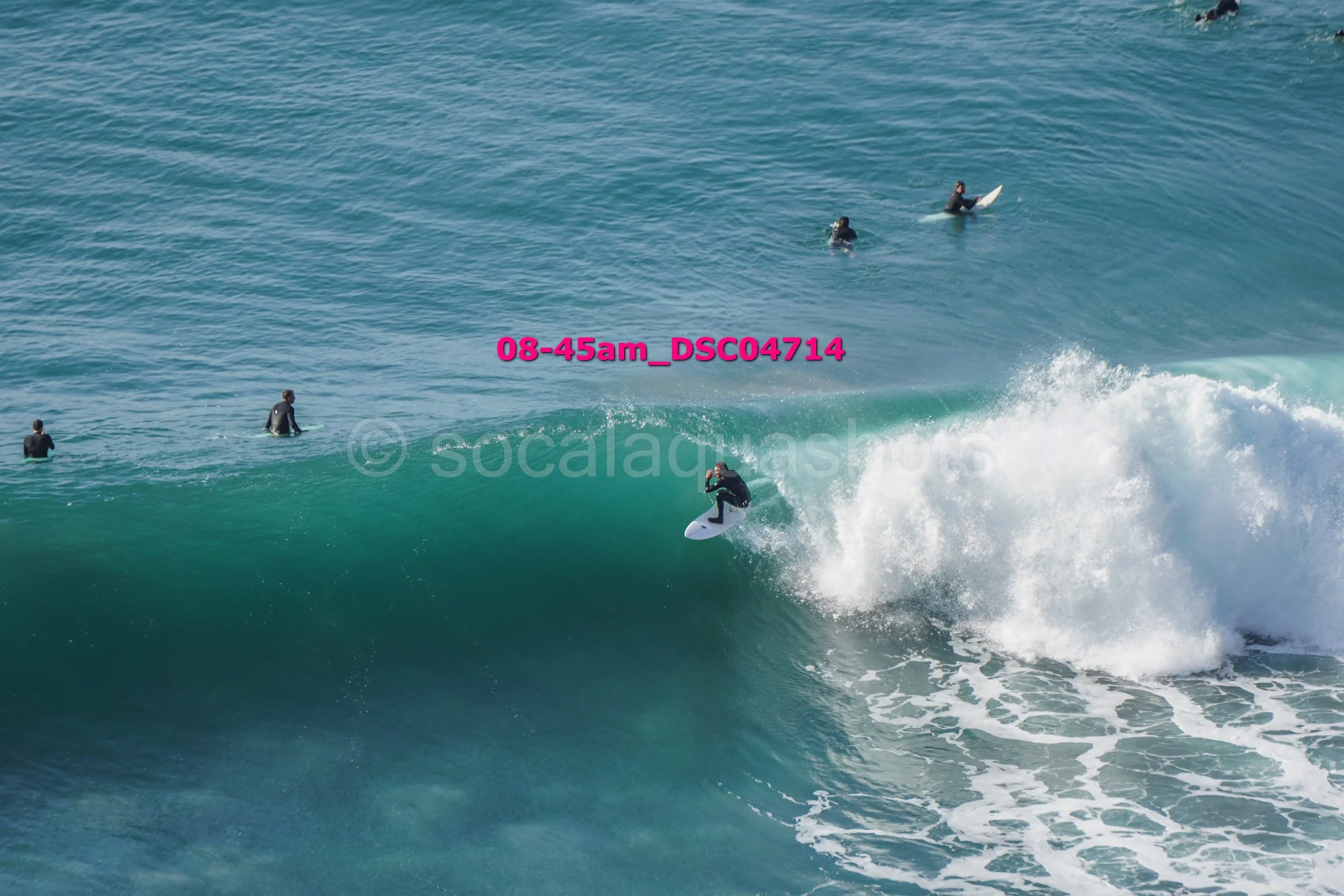 Surfer riding a large wave with five people in the water watching in the background.