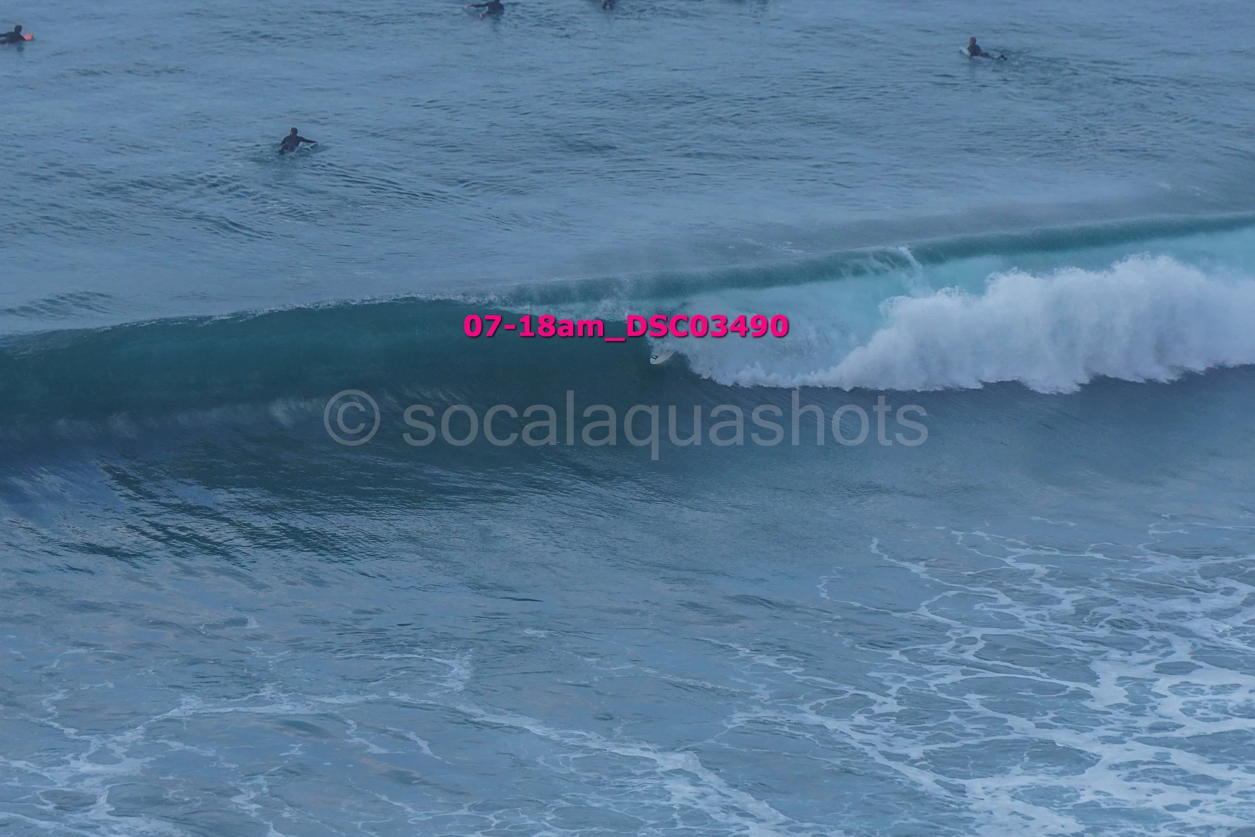 A surfer riding a wave in the ocean with several other surfers visible in the water behind.