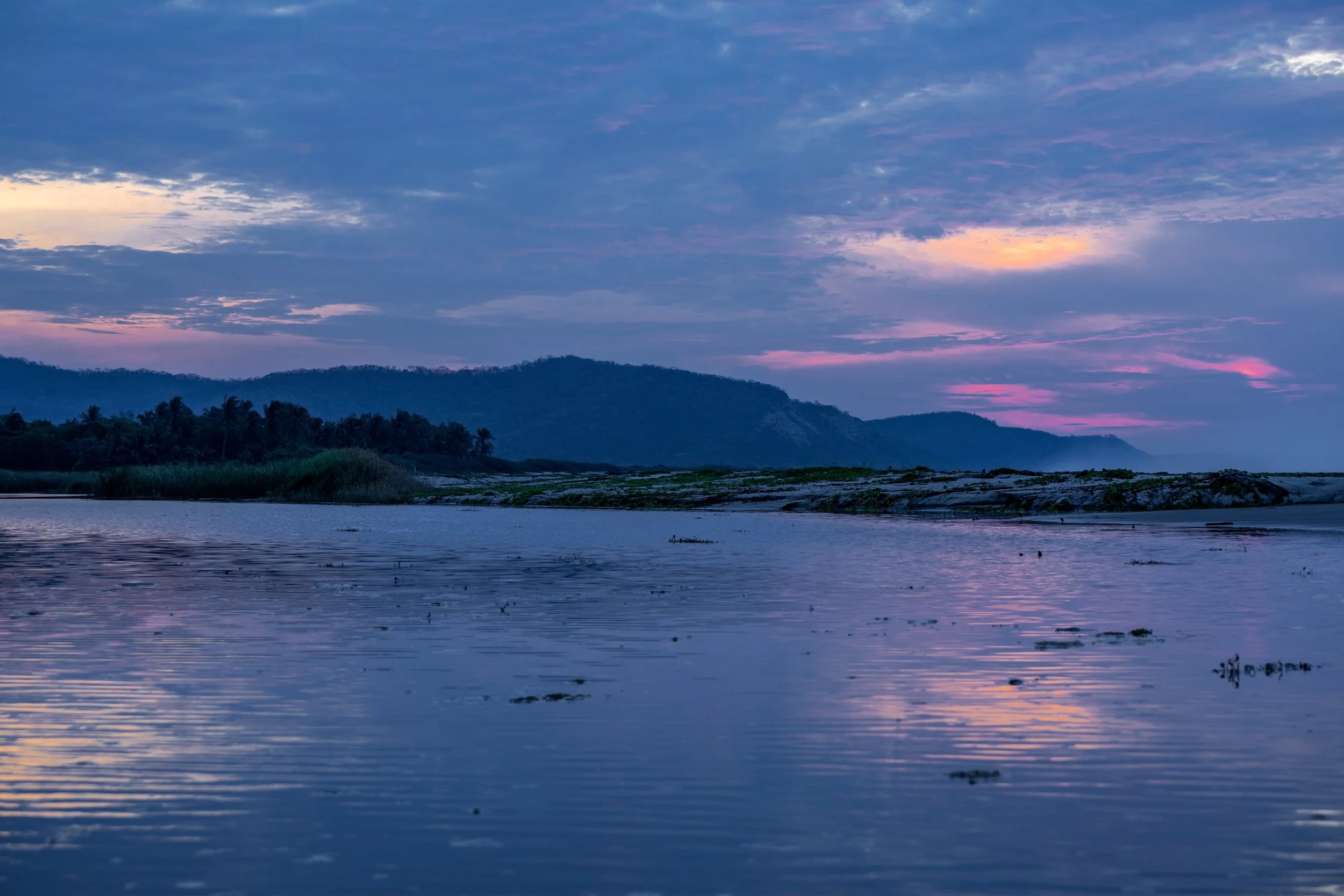 A scenic view of a river at sunset with a mountain range in the background and purple and pink clouds in the sky.