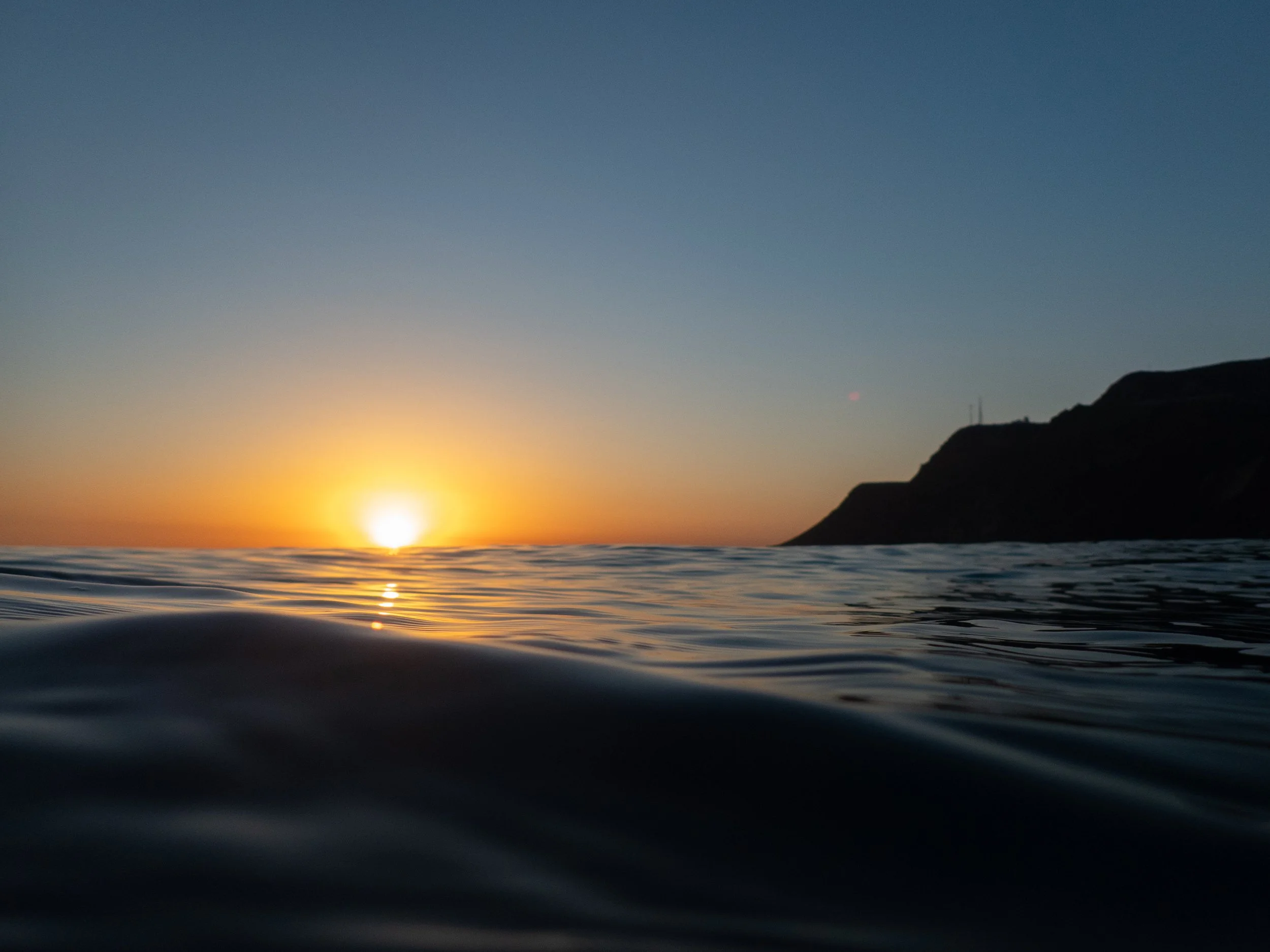 Sunset over the ocean with a mountainous coastline in the distance.