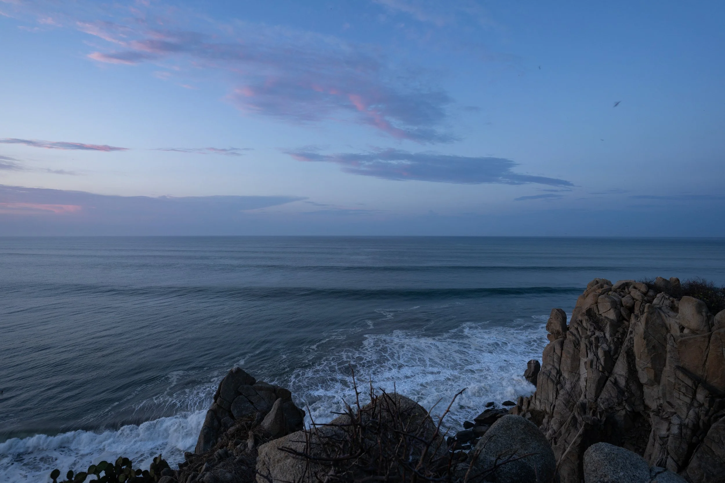 Seascape with calm ocean water, rocks along the shoreline, and a sky with scattered clouds at dusk or dawn.