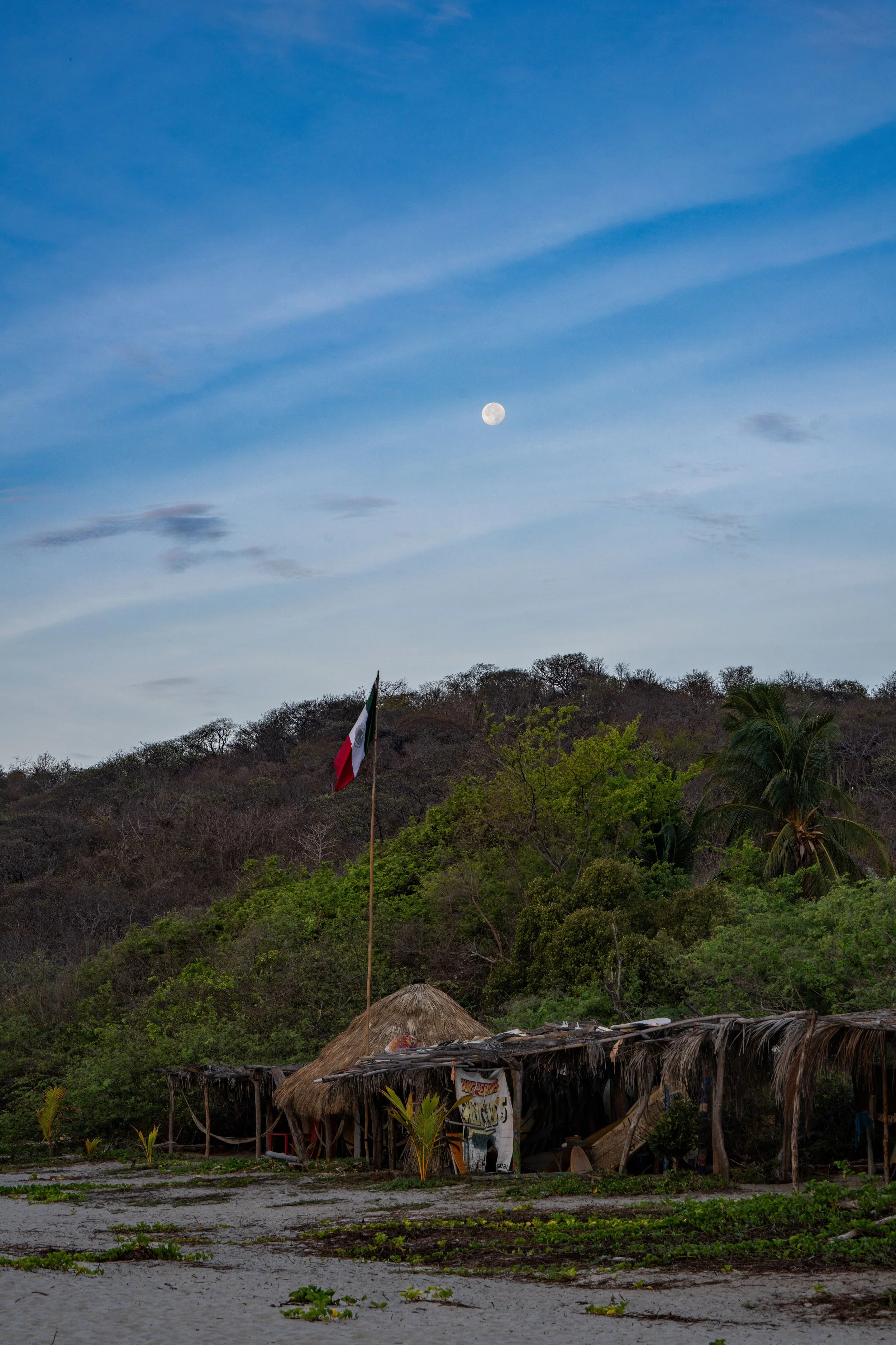 A rural scene showing a small thatched hut with a Mexican flag on a pole, set against a hillside with trees. The sky is partly cloudy with the moon visible during dusk.