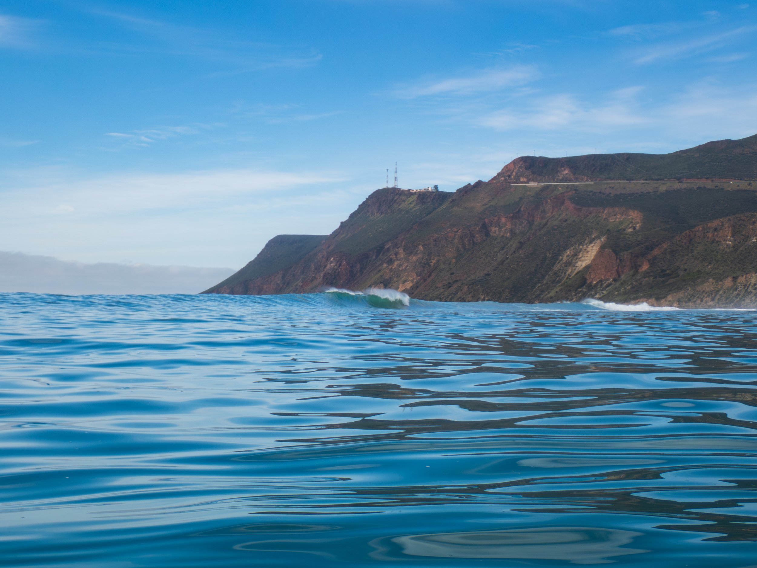 View of the ocean with small waves and a mountainous coastline in the background under a partly cloudy sky.