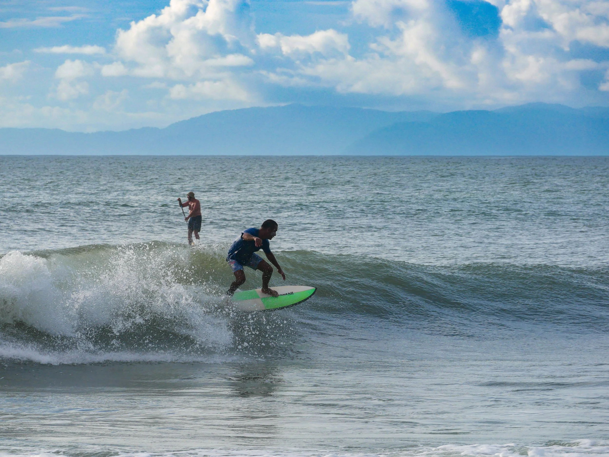 Person surfing on a wave and another paddleboarding in the ocean under a cloudy sky.