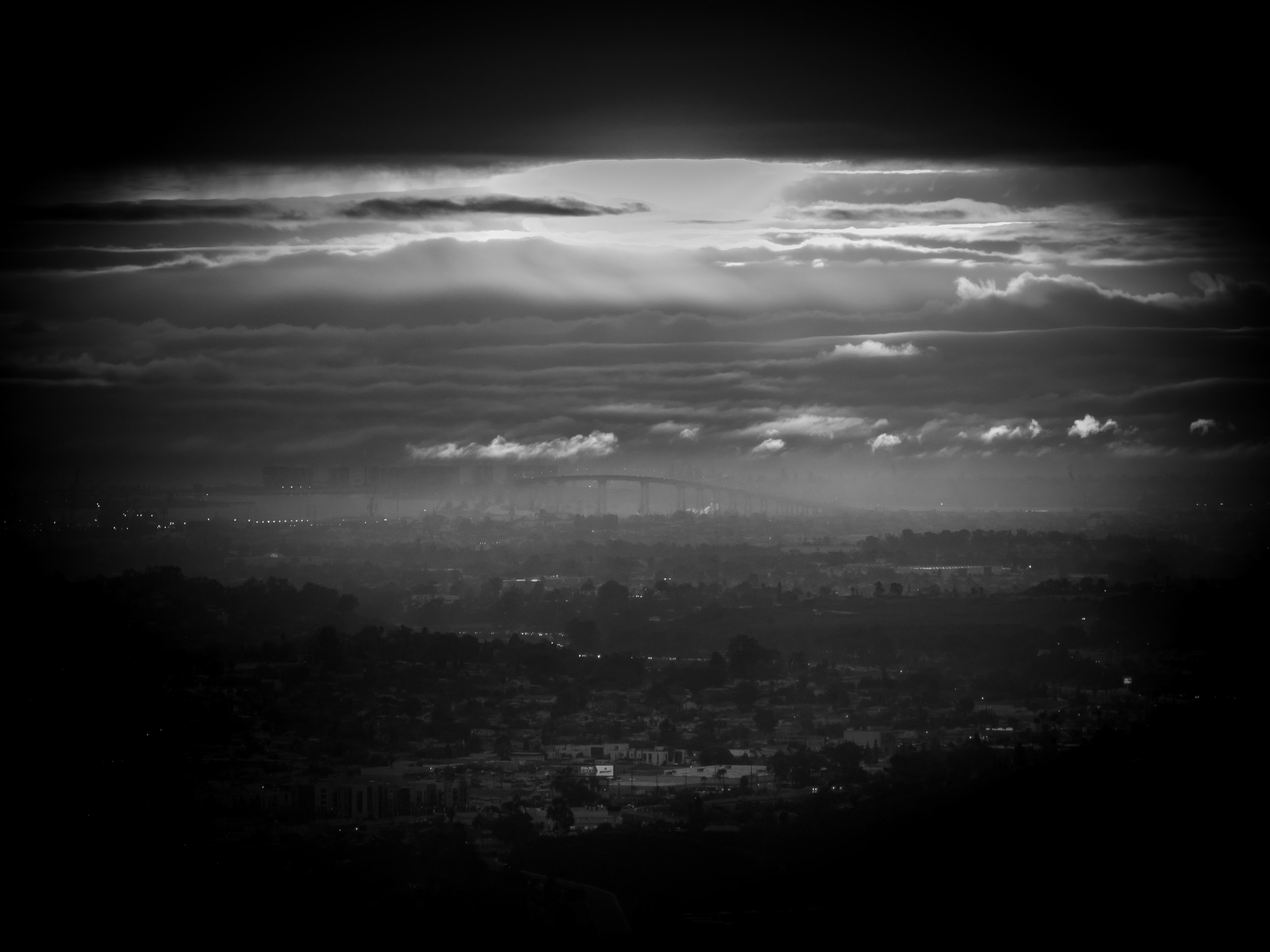 Black and white photograph of a cityscape with scattered buildings, a distant bridge, and a dramatic cloudy sky with rays of sunlight breaking through.