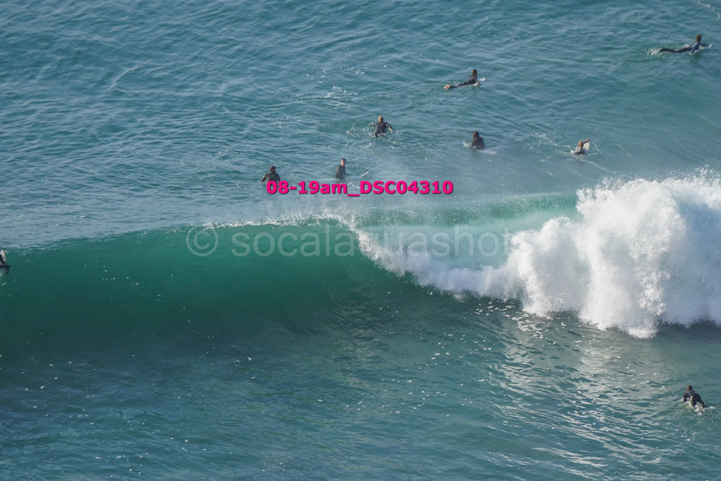 Several surfers riding a large wave in the ocean, some waiting for their turn and others paddling out.