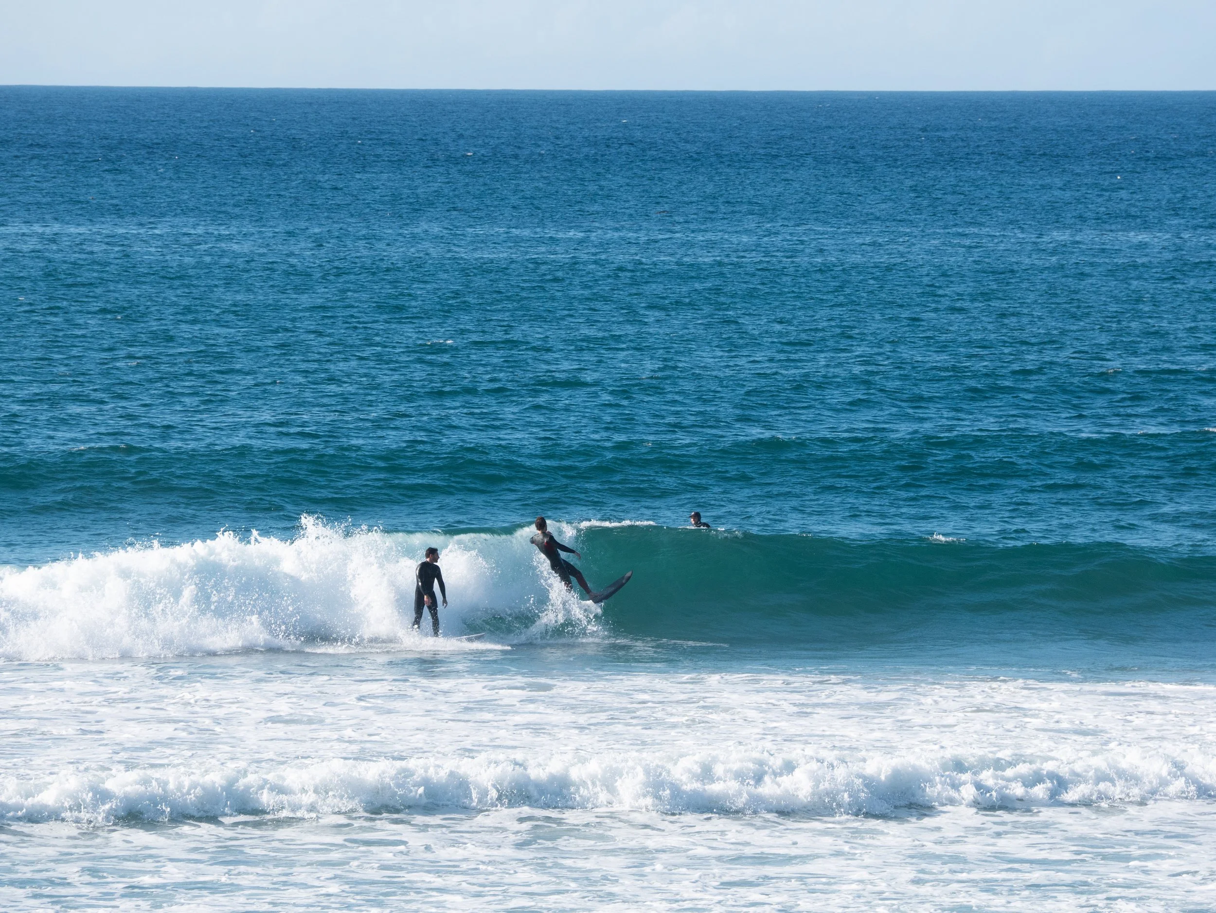 Three surfers in wetsuits riding and waiting for waves in the ocean, with a clear blue sky overhead.