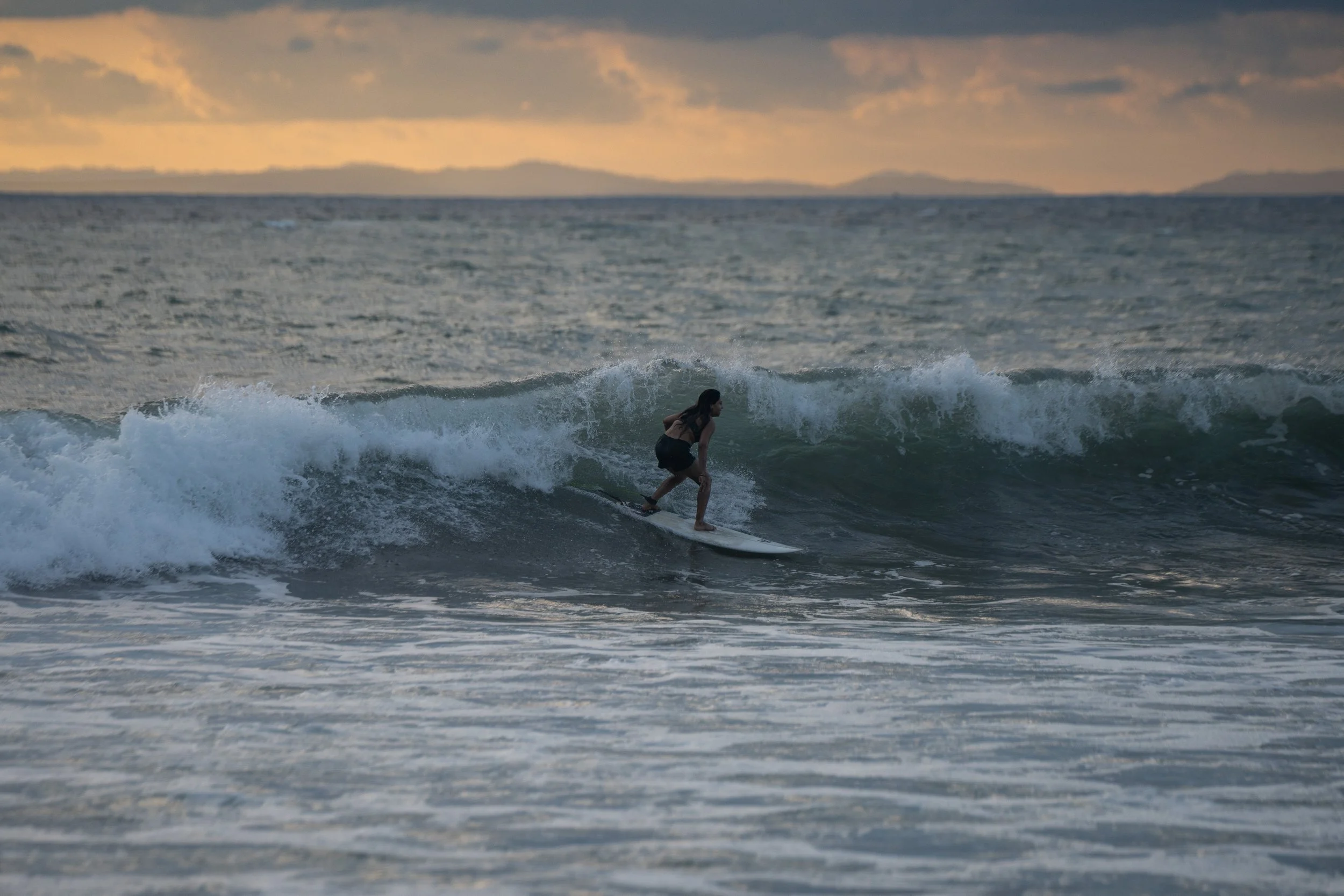 A woman surfing on a wave at sunset with a distant shoreline and cloudy sky in the background.