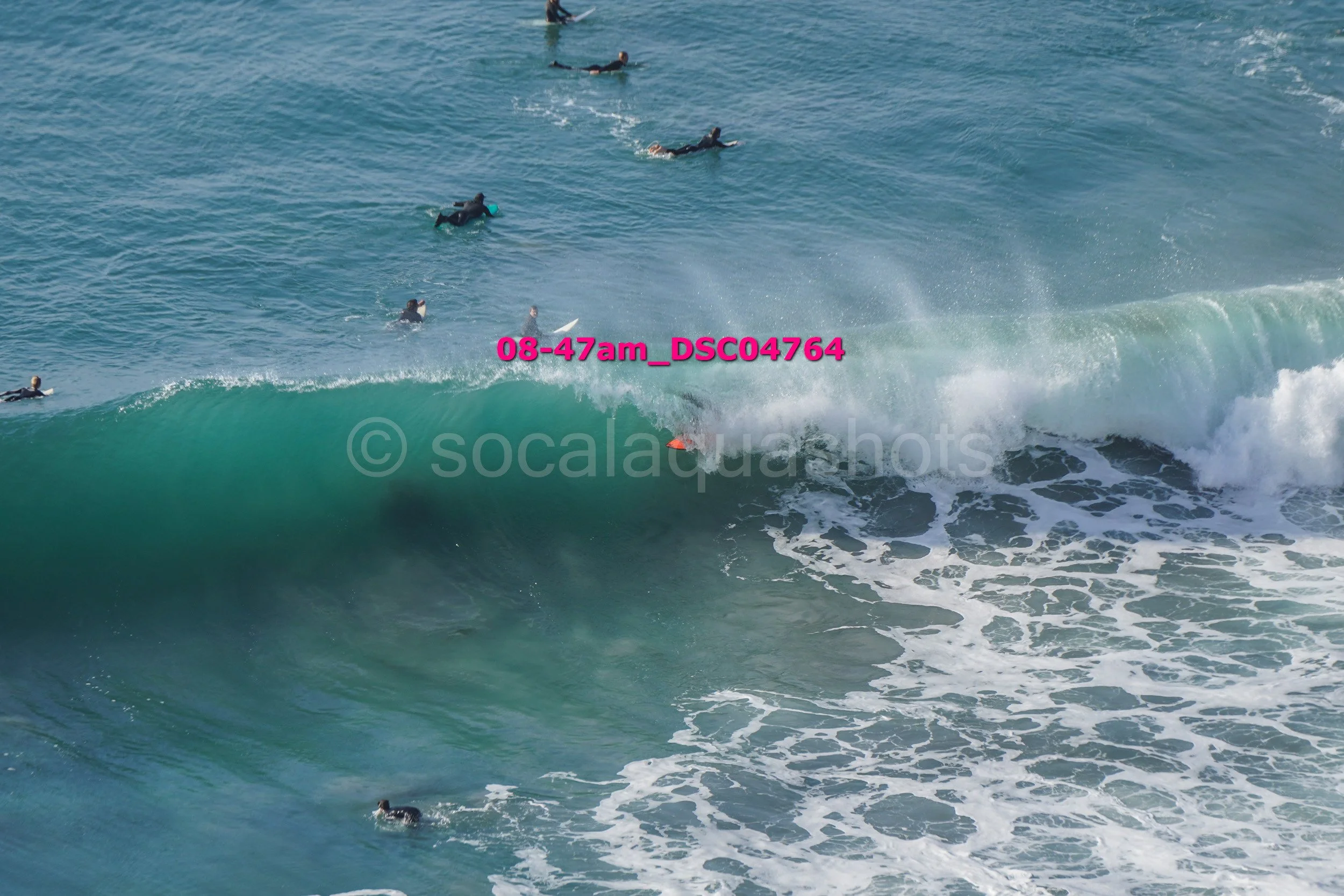 Surfers waiting in the water to catch a wave, with one surfer riding a large wave close to the shore.