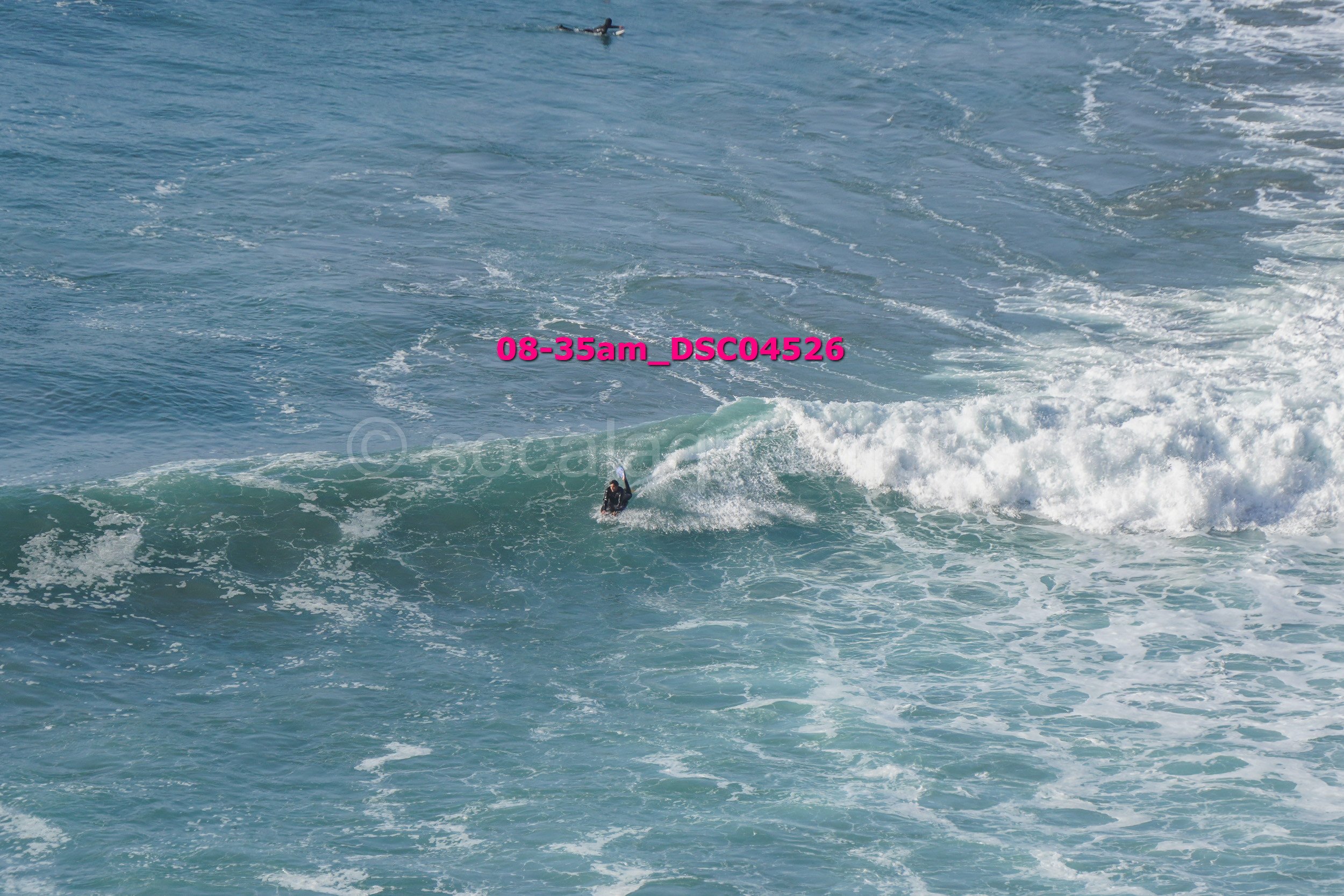 A person surfing on a wave in the ocean, with another surfer visible farther back.