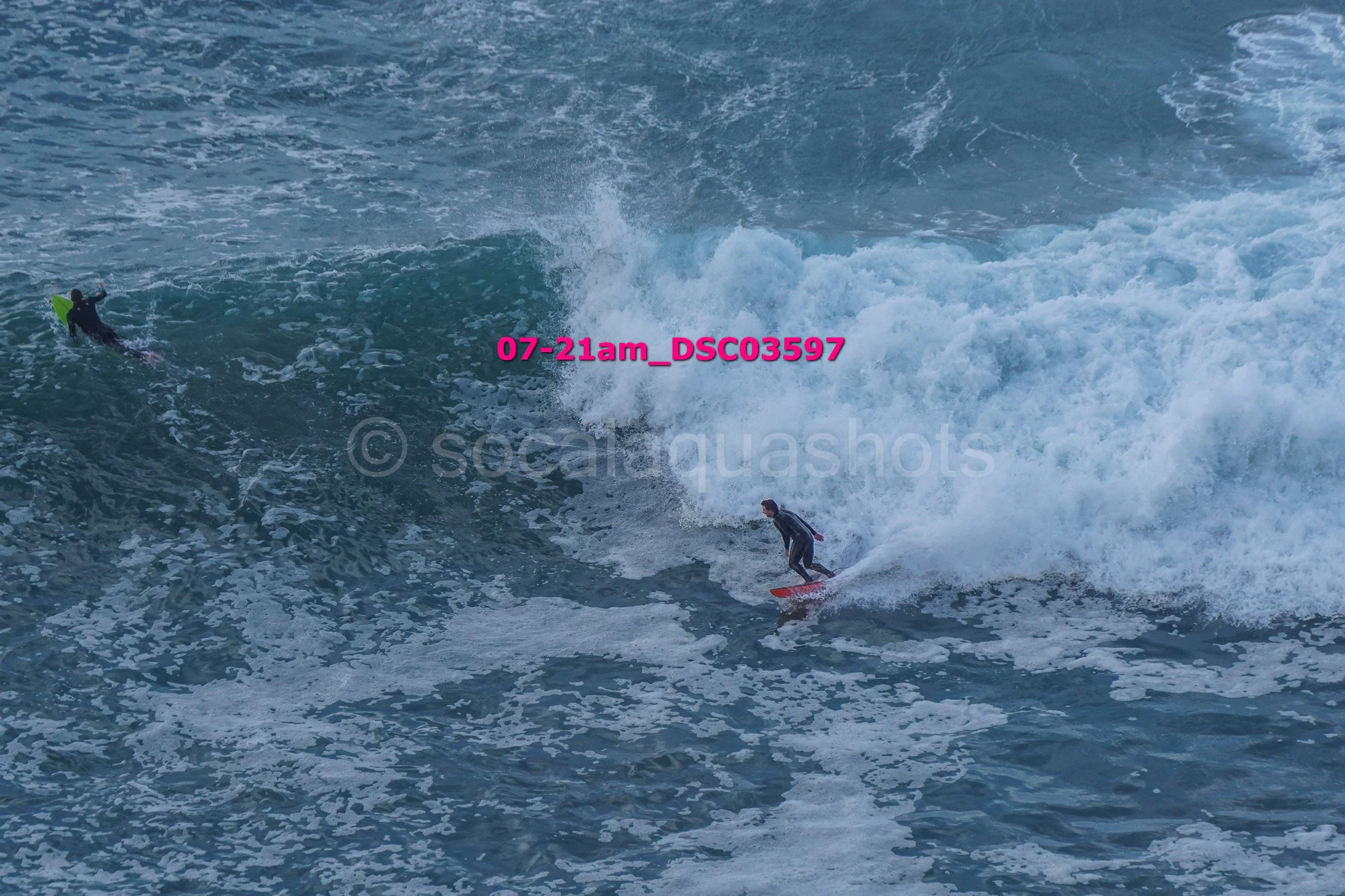 Two surfers riding large ocean waves, with one surfer closer to the camera and the other further back; ocean with white foam and crashing waves.