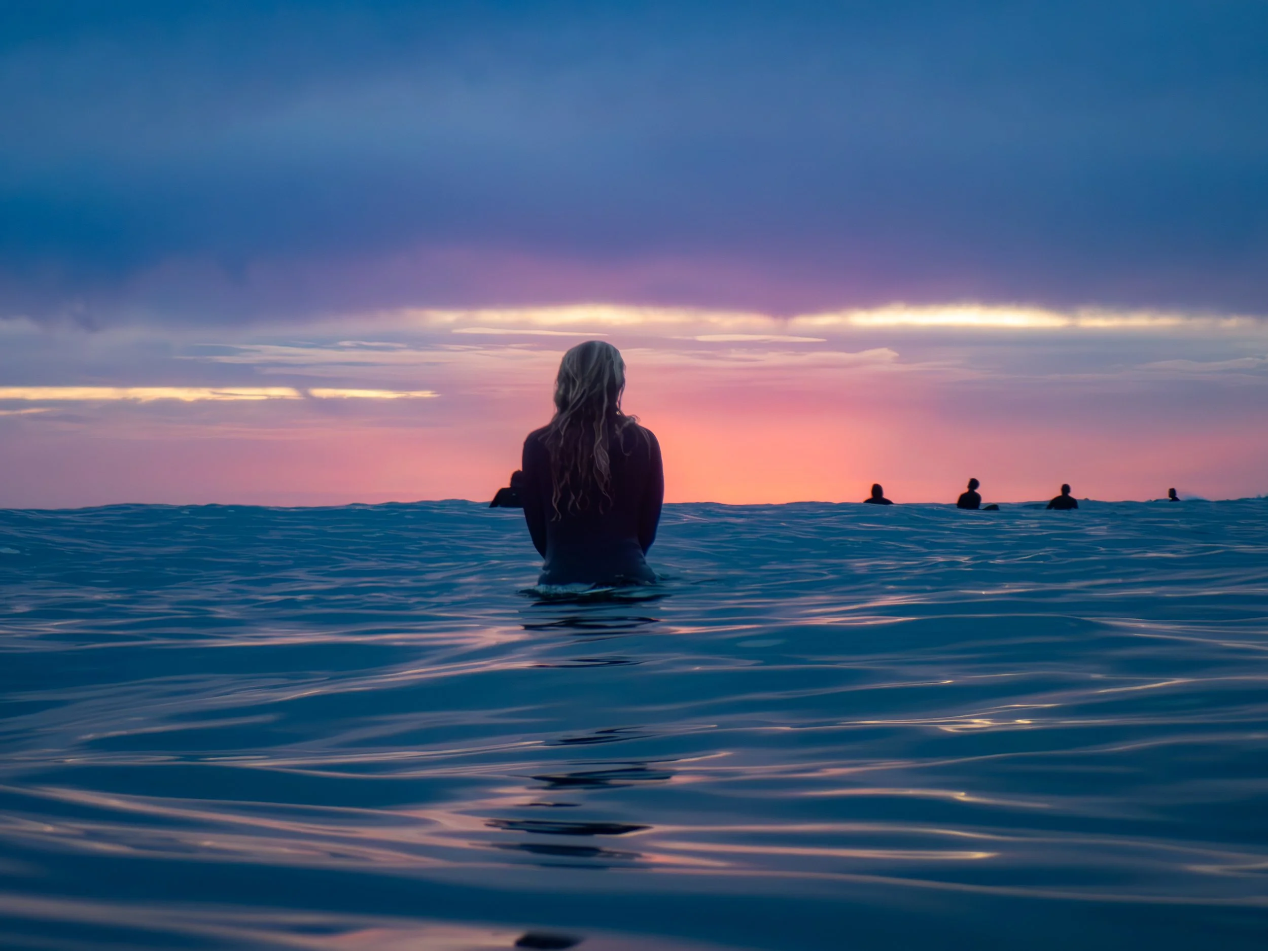 A woman stands in the ocean facing the sunset, with a group of people in the distance. The sky has shades of pink, purple, and blue with clouds.