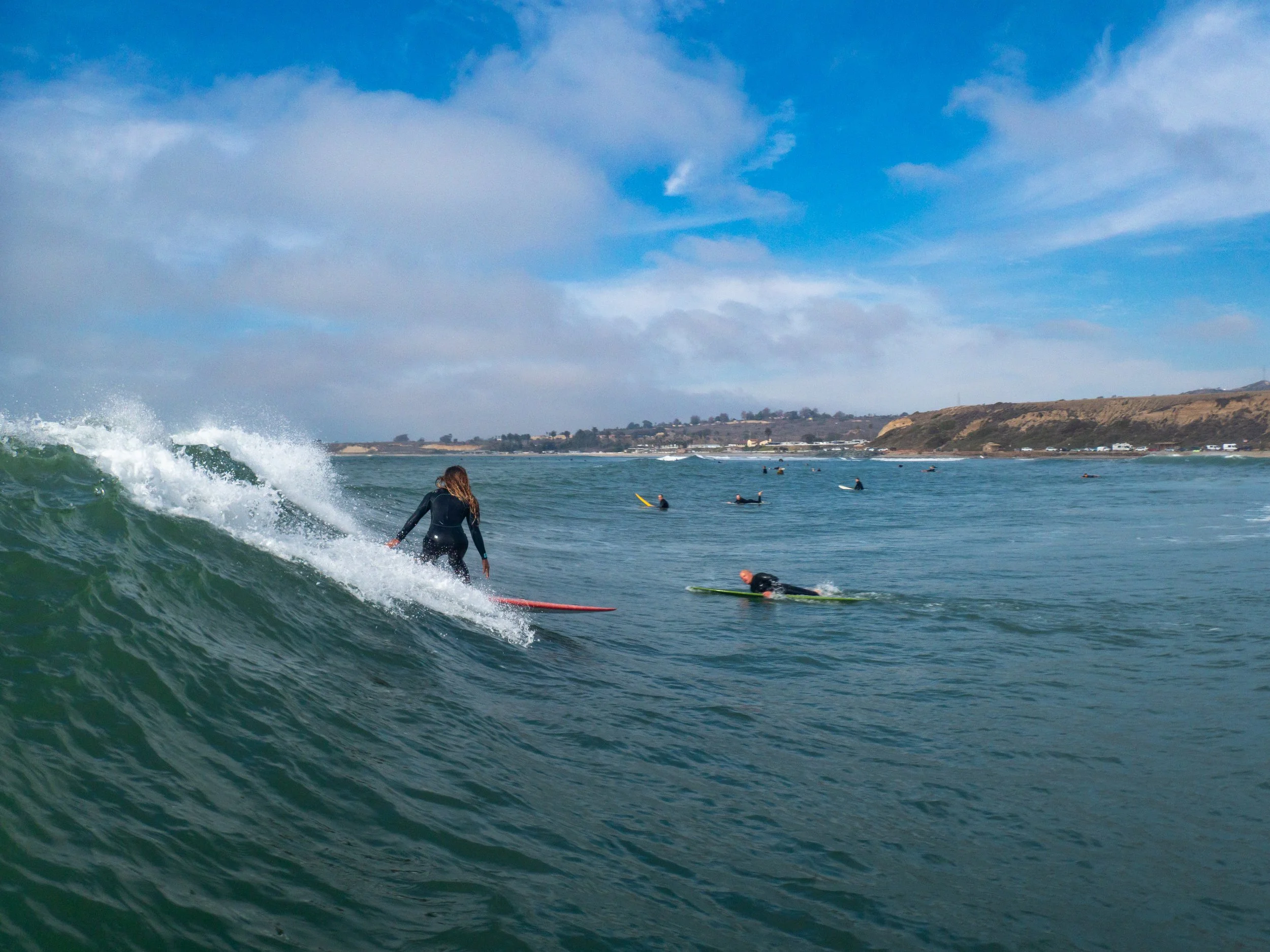 A woman surfing on a wave in the ocean with several other surfers in the background, coastline visible under partly cloudy sky.