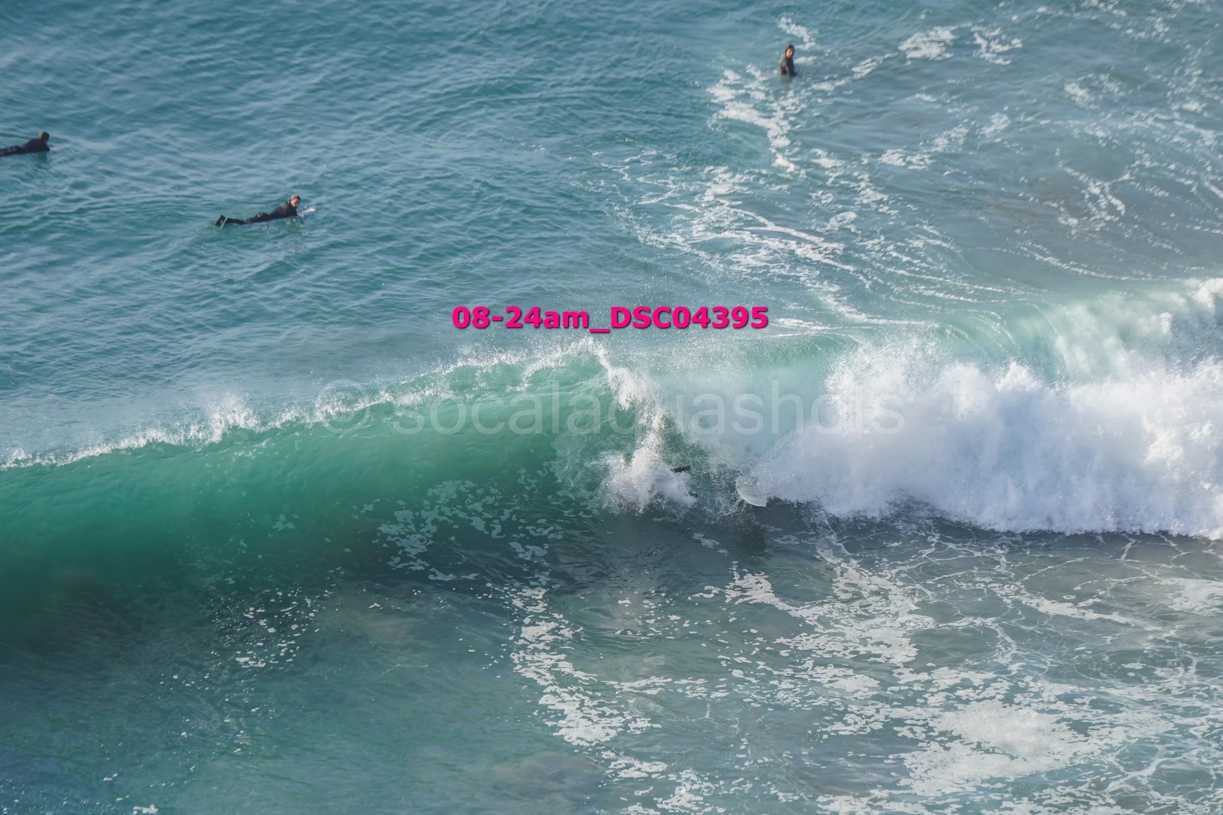 People in wetsuits surfing and swimming in the ocean, with a large wave crashing and a surfer riding the wave.