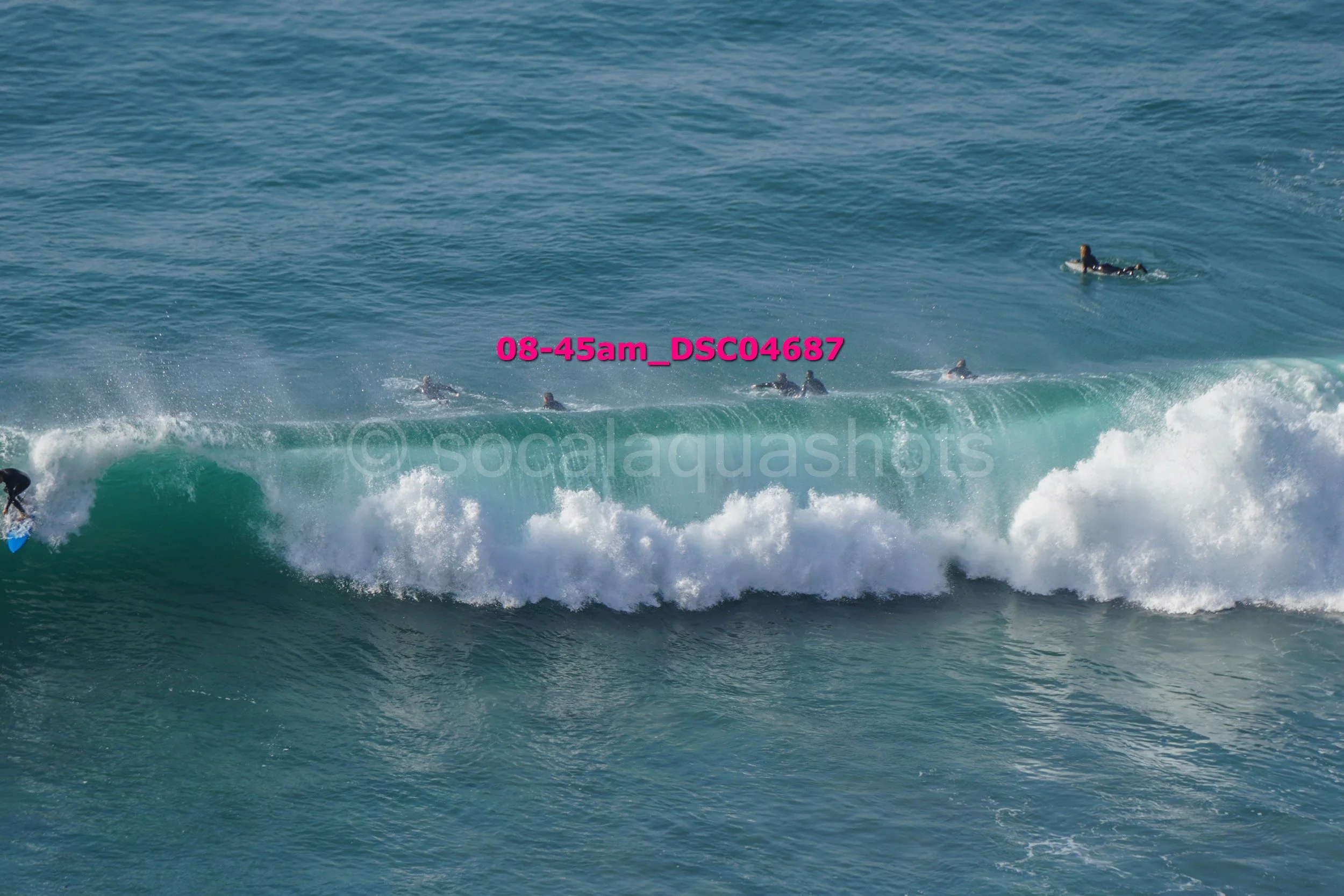 Surfers riding a wave in the ocean. One surfer on the left with a blue surfboard, others in the water, and a large breaking wave.