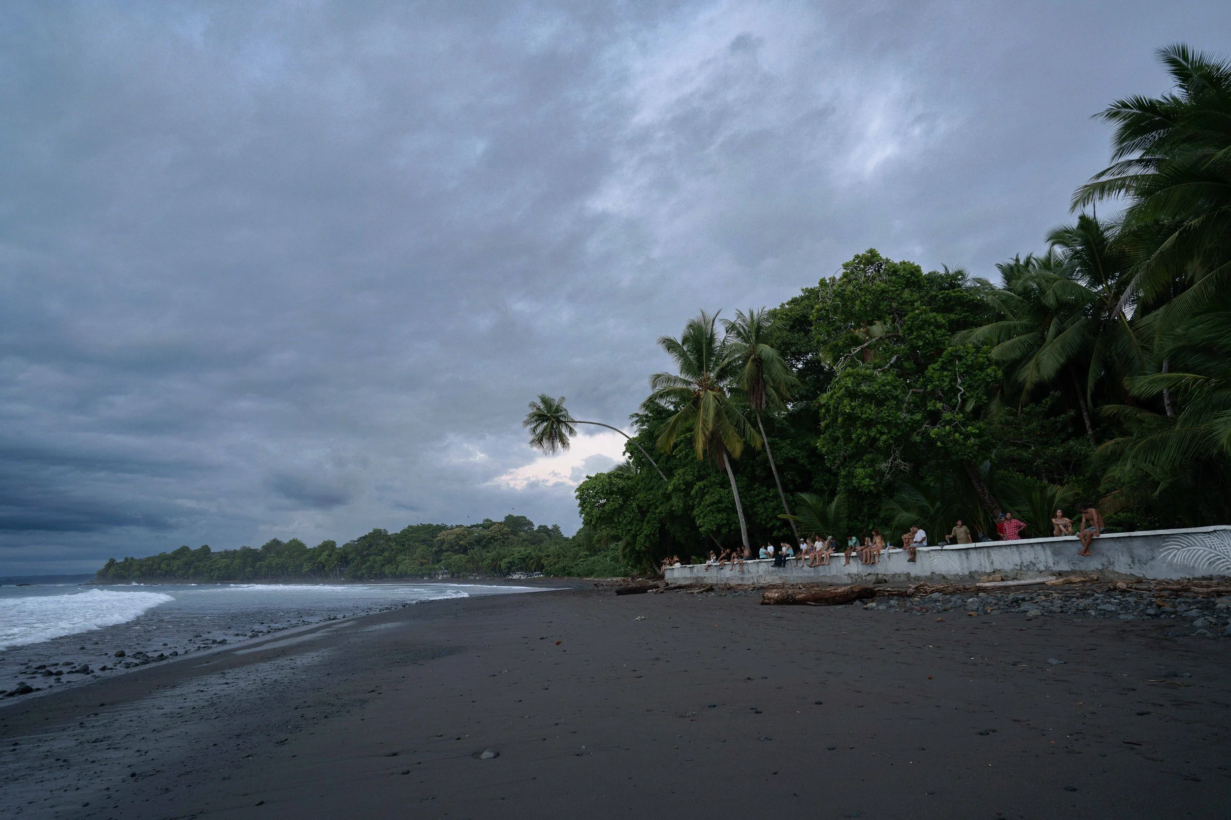Overcast sky over a beach with a row of people sitting and standing along a white wall, tropical trees including palm trees, ocean waves, and a distant shoreline.