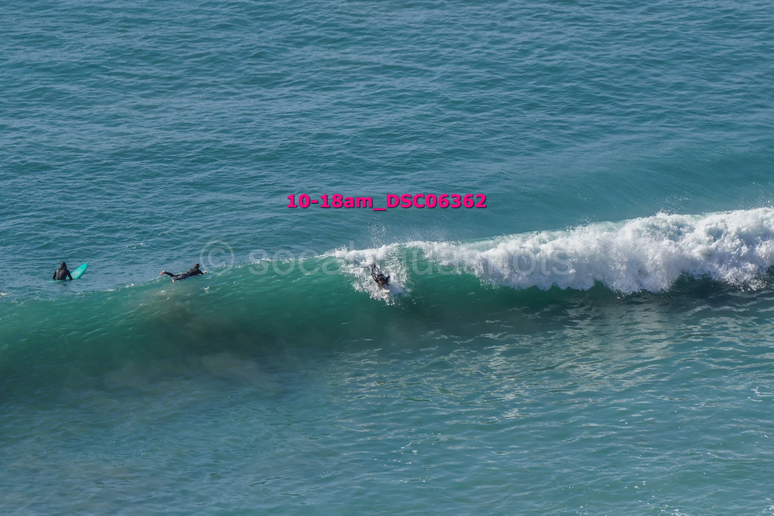 Two surfers in wetsuits riding a wave, with one lying on their surfboard and the other standing, in the ocean during daylight.