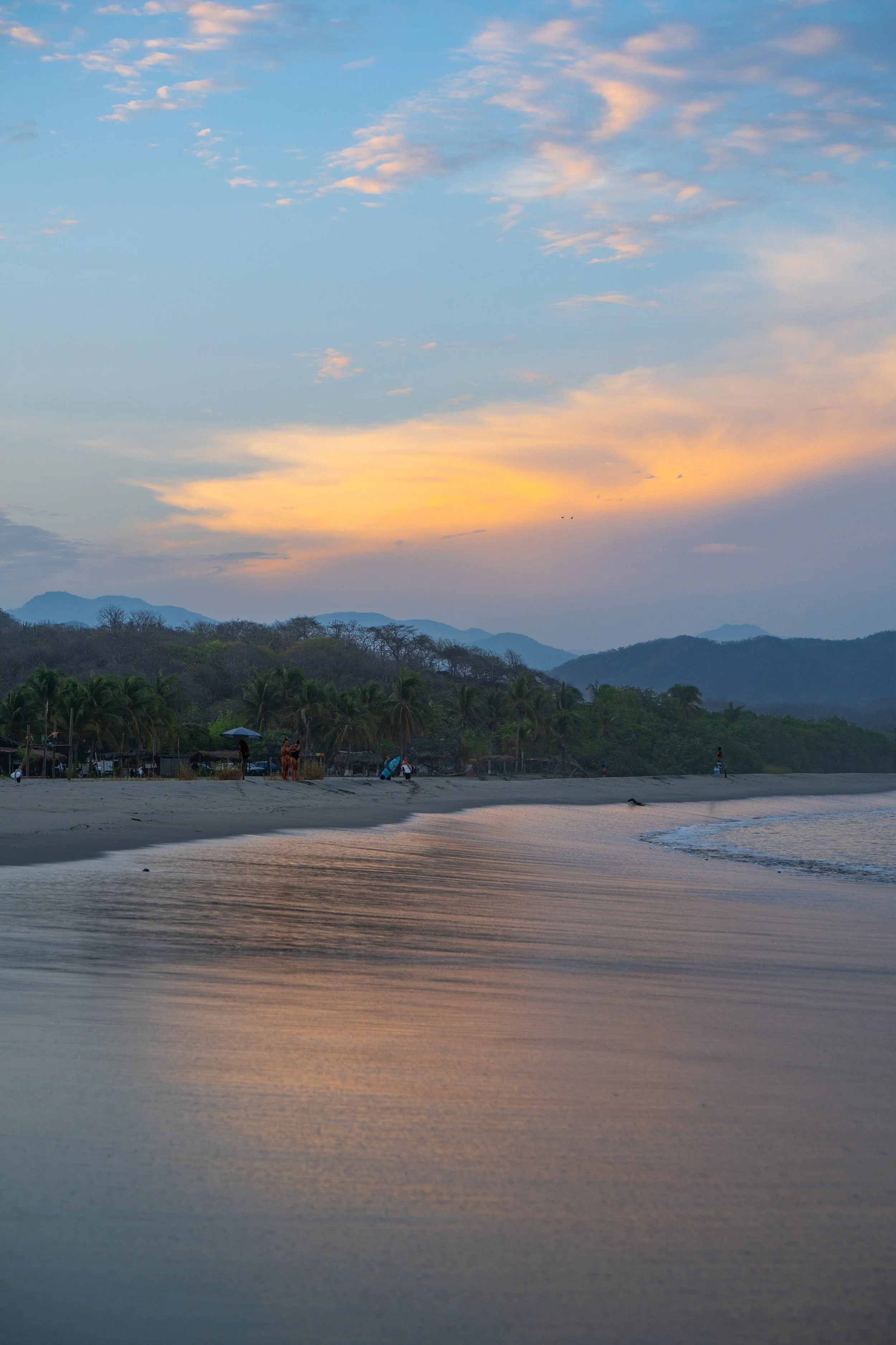 Sunset over a beach with calm water, distant mountains, and a cloudy sky.