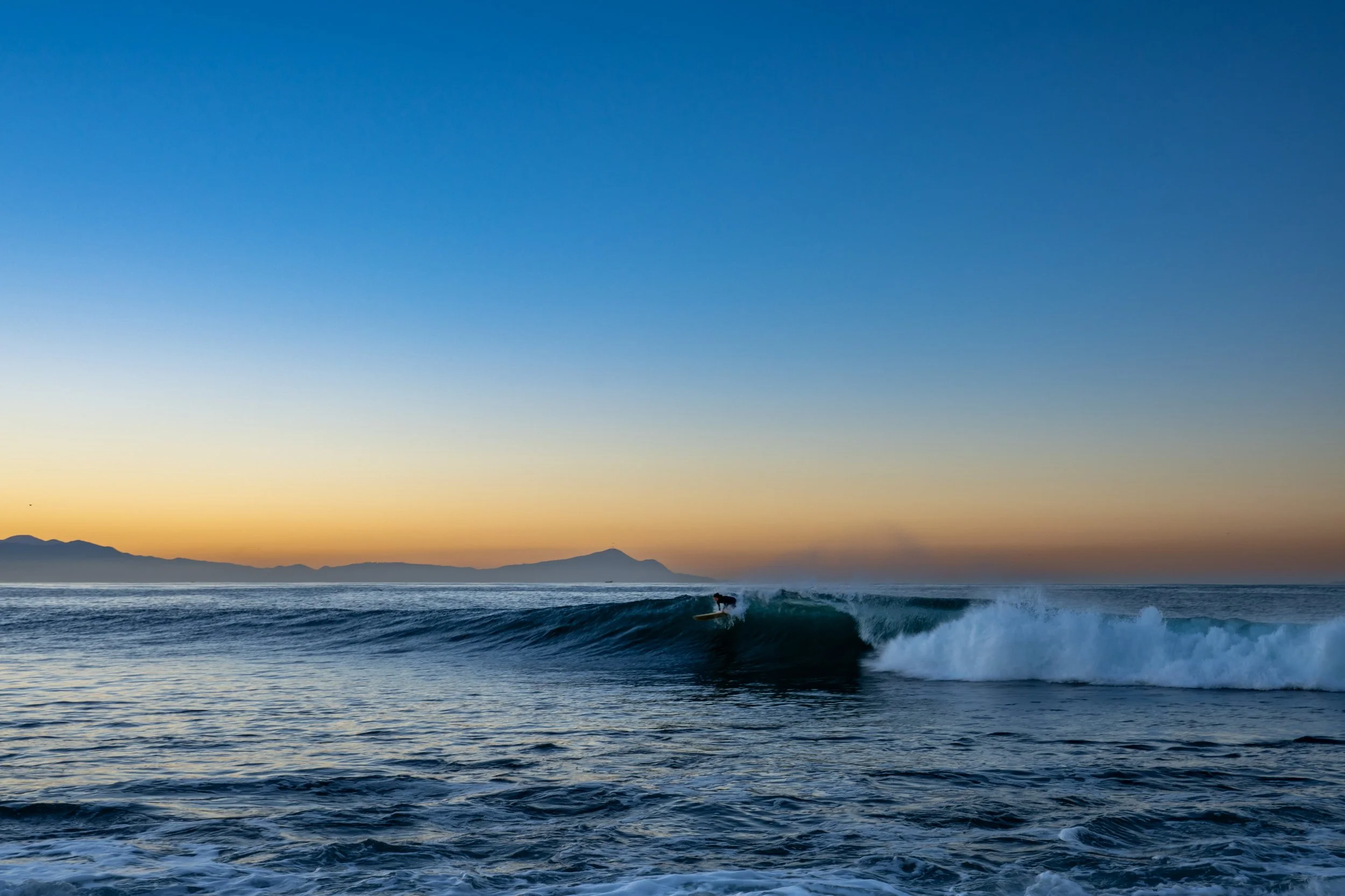 A person surfing on a wave in the ocean during sunset with mountains in the background.