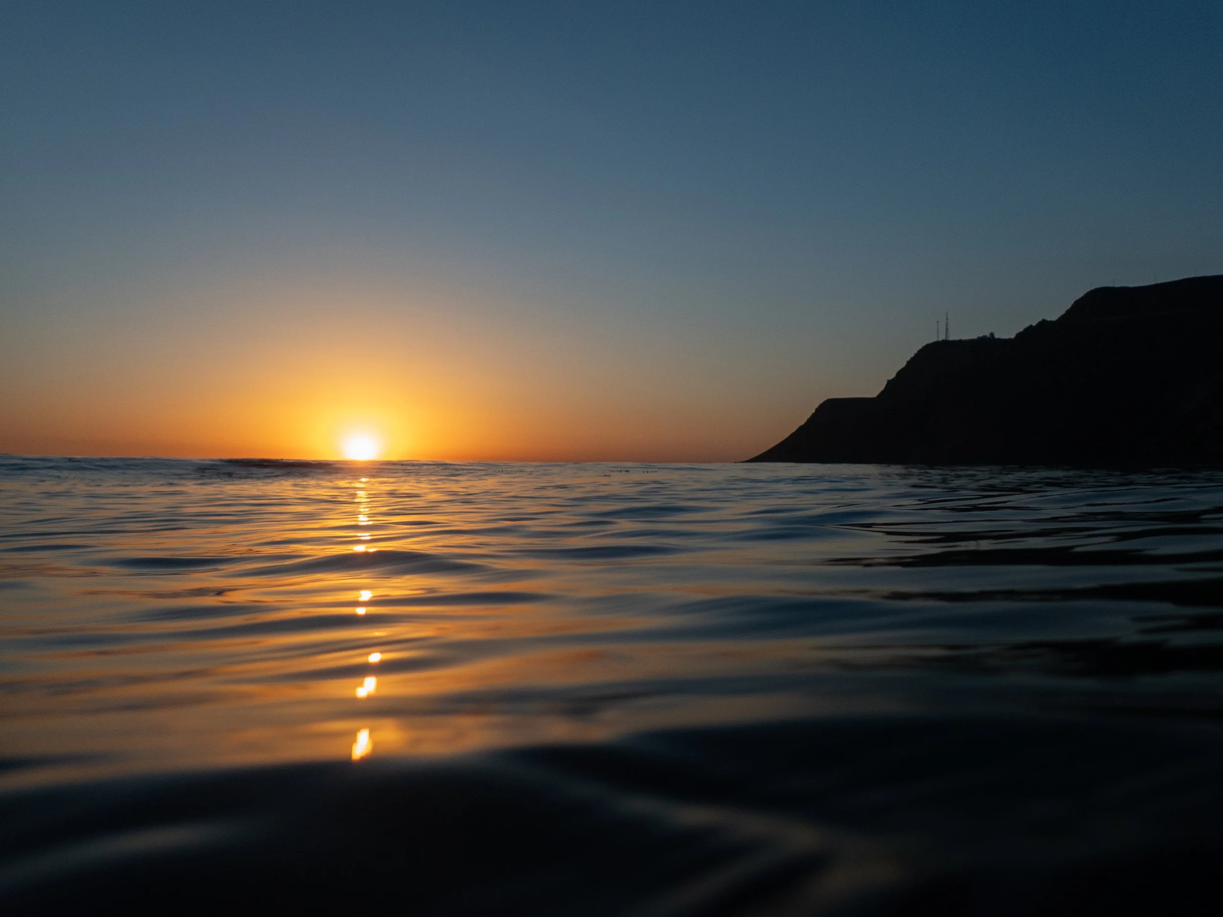 Sunset over the ocean with a silhouette of a cliff on the right and calm water reflecting the orange and yellow glow of the sunset.