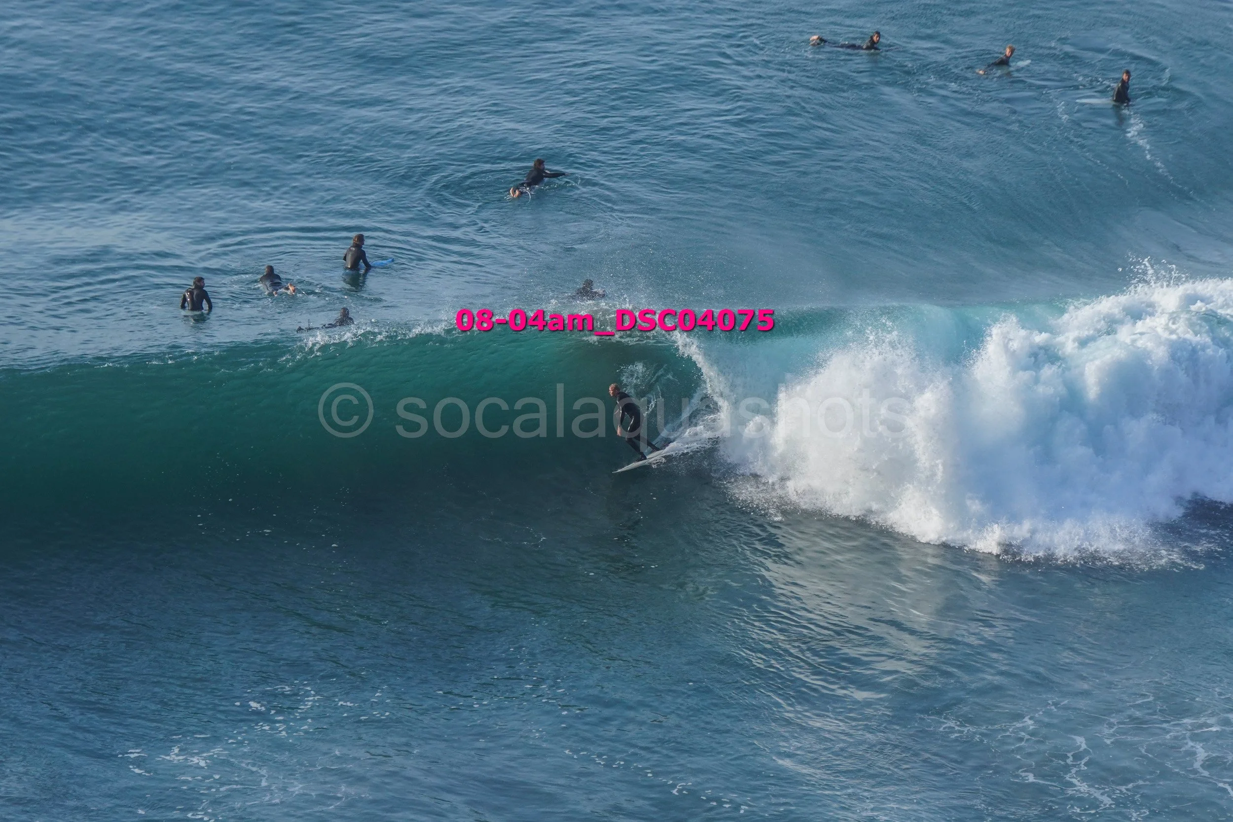 A surfer riding a wave while multiple people, dressed in wetsuits, watch from the water.