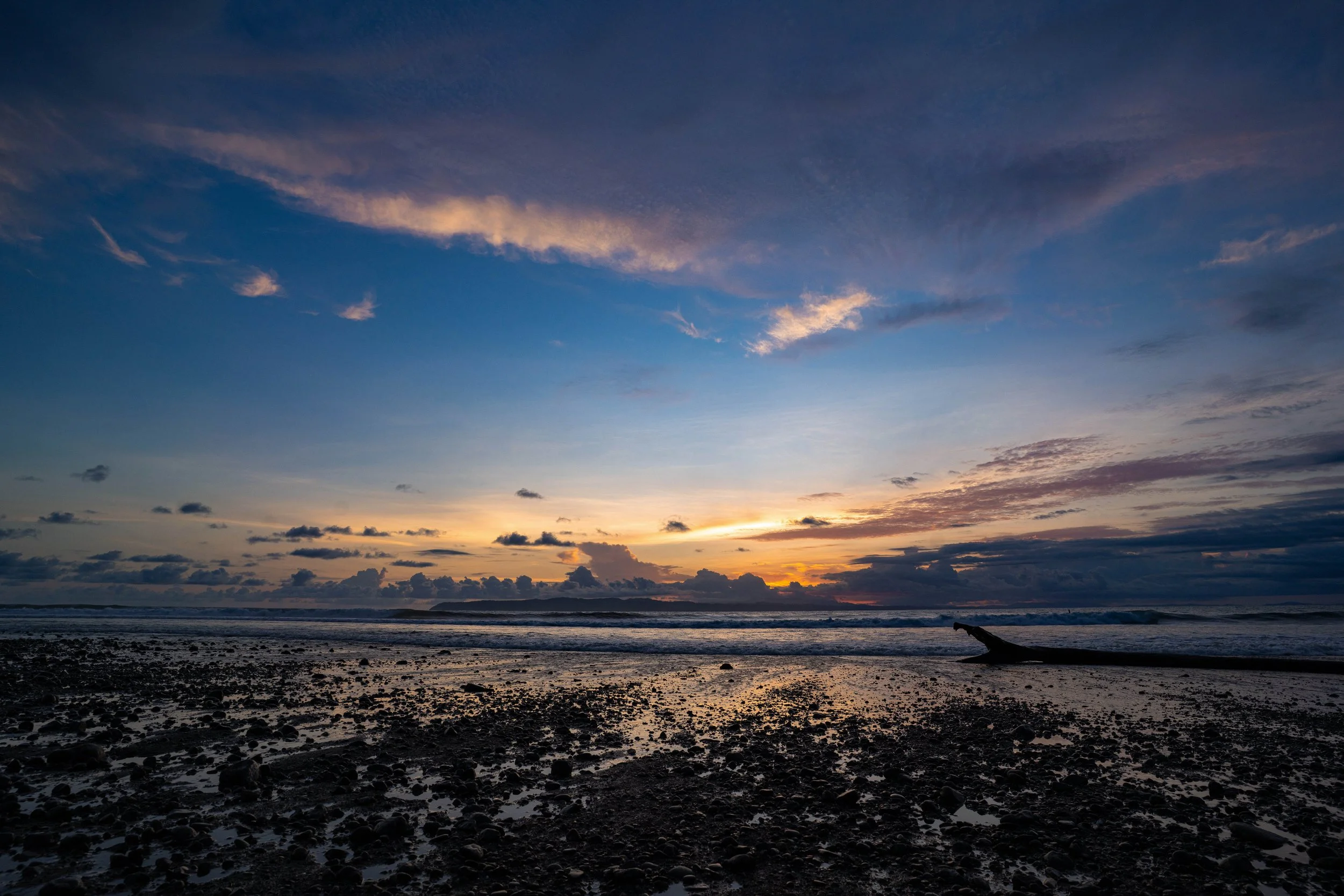 A beach shoreline at sunset with dark rocks and reflective water, a distant horizon, and a partly cloudy sky with soft clouds illuminated by the setting sun.