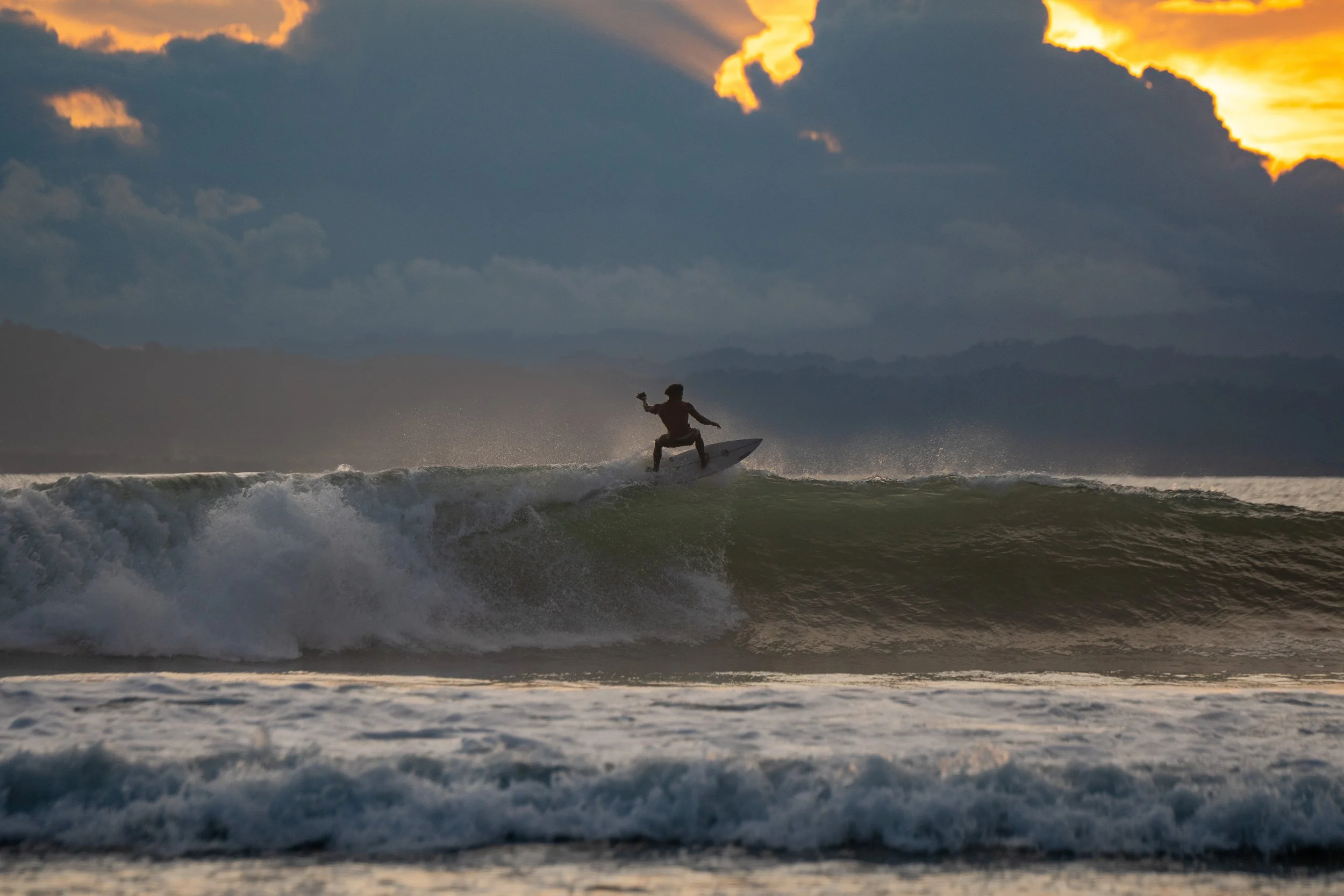 A person surfing on a wave during sunset with cloudy sky and distant mountains in the background.