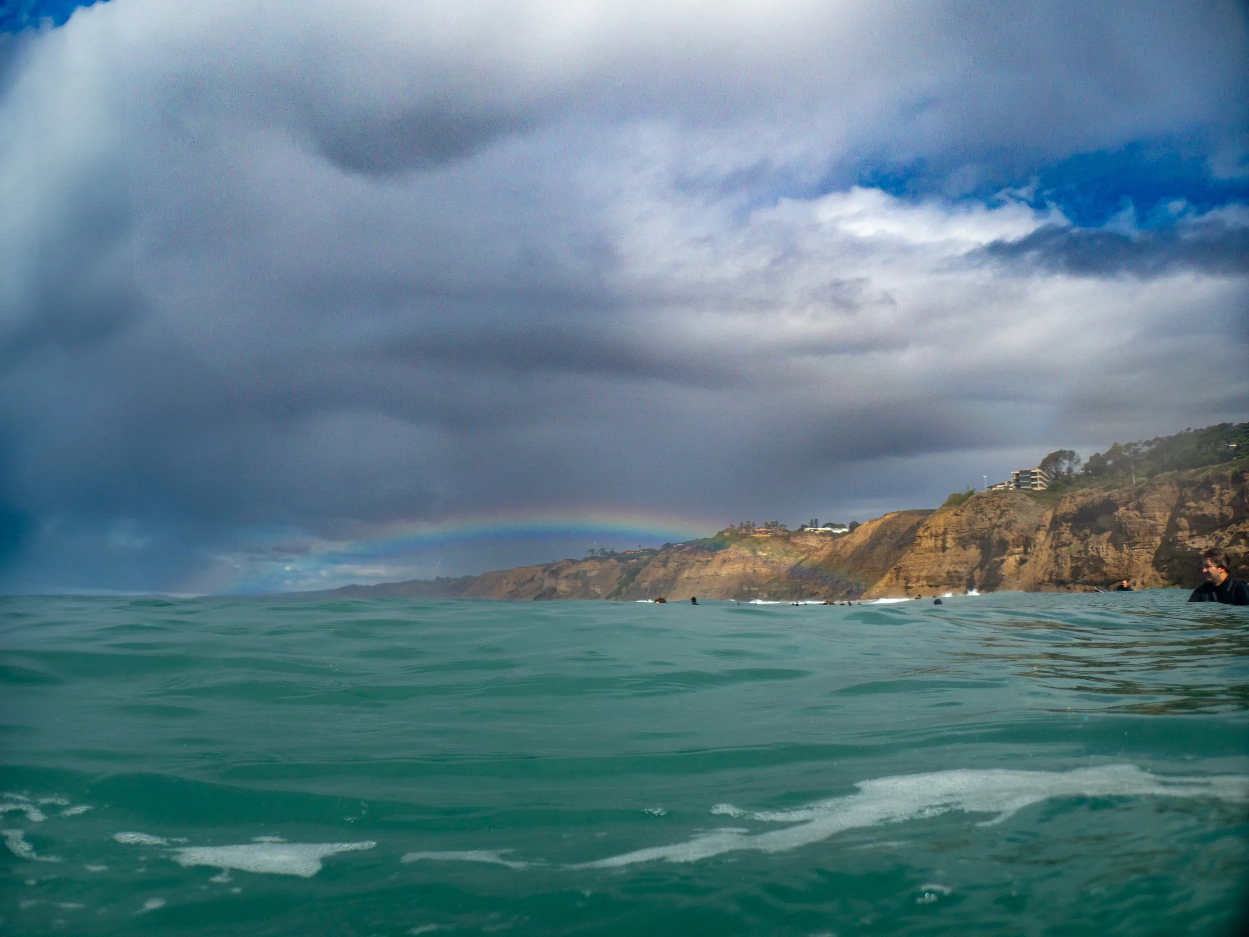 Ocean with teal waves and cliffs in the background under a sky with dark clouds and a faint rainbow.
