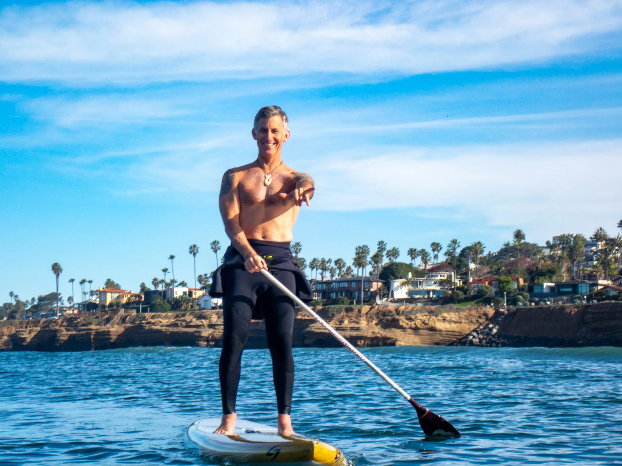 A shirtless man paddleboarding on the water, smiling and pointing towards the camera, with a coastline and palm trees in the background.