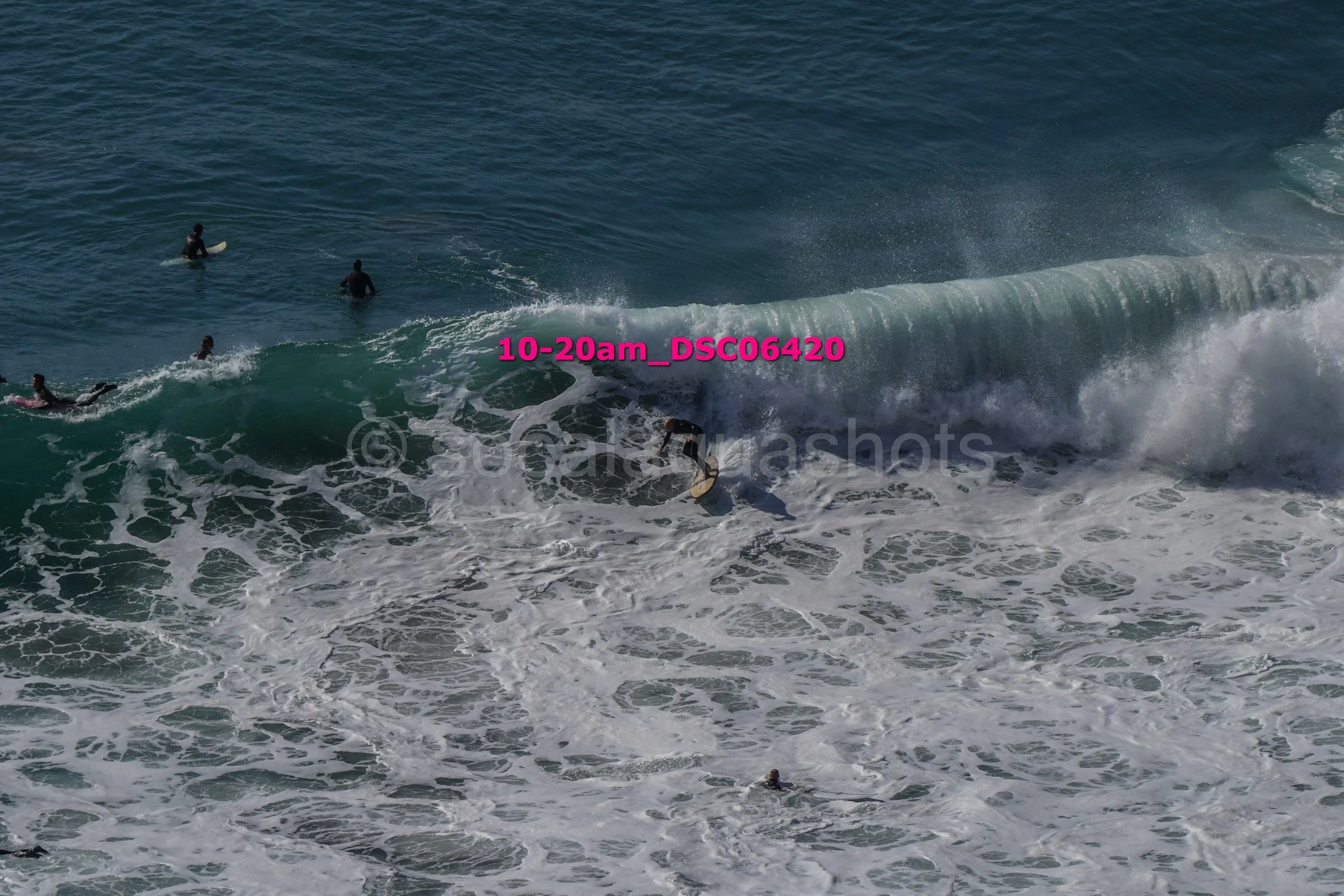Surfer riding a large wave with several other surfers in the water nearby.