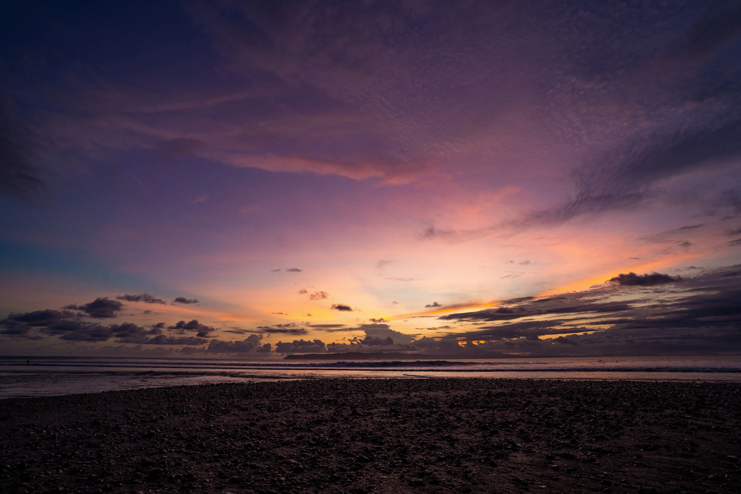 A sunset over the ocean with a colorful sky and scattered clouds, viewed from a rocky beach.