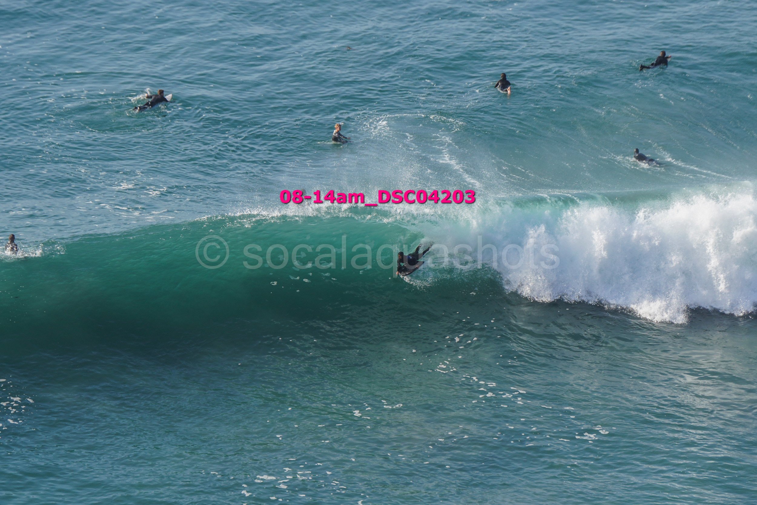 Surfer riding a wave with several people in the water watching from above.