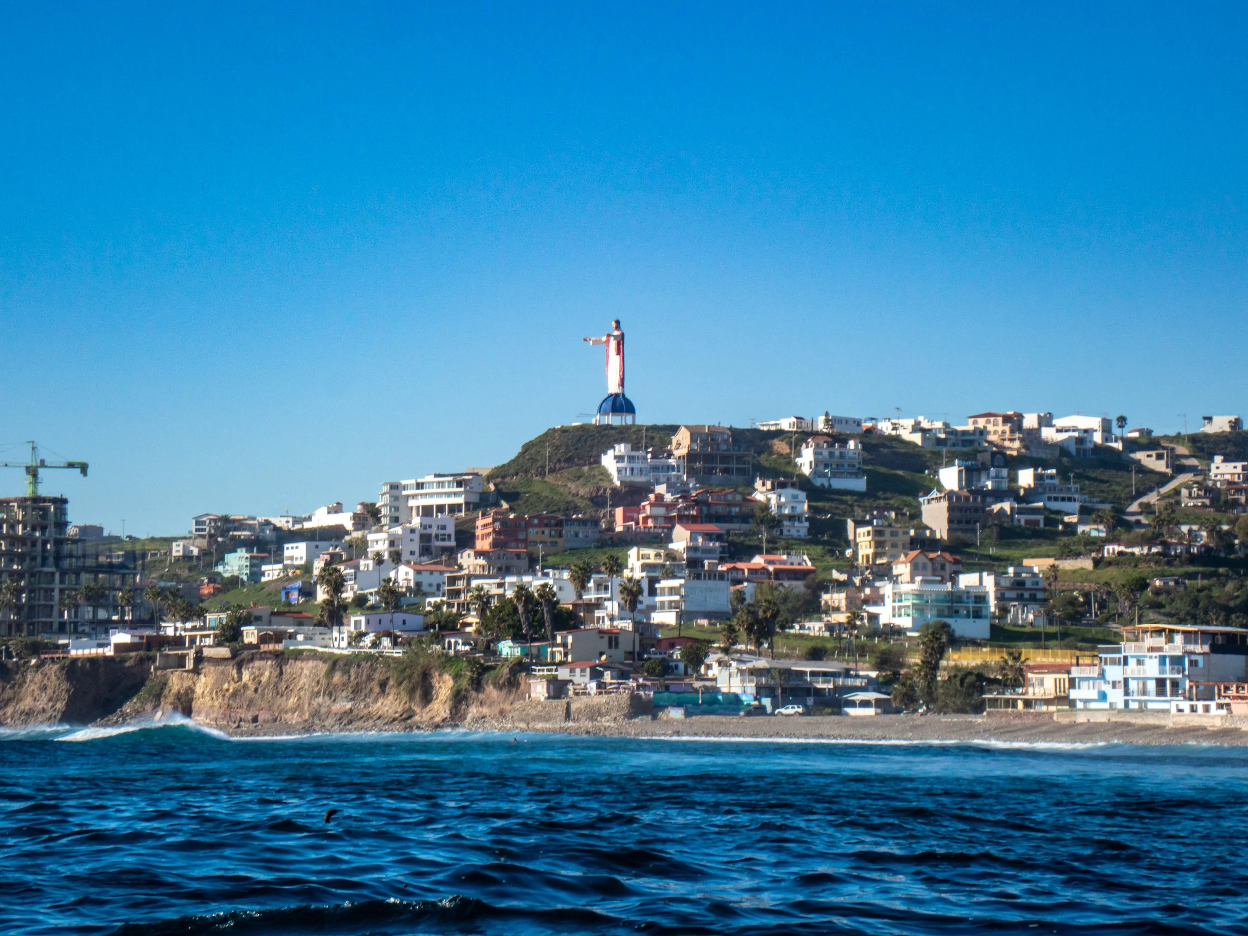 Coastal hillside with numerous houses and a Christian cross statue on top, overlooking the ocean under a clear blue sky.