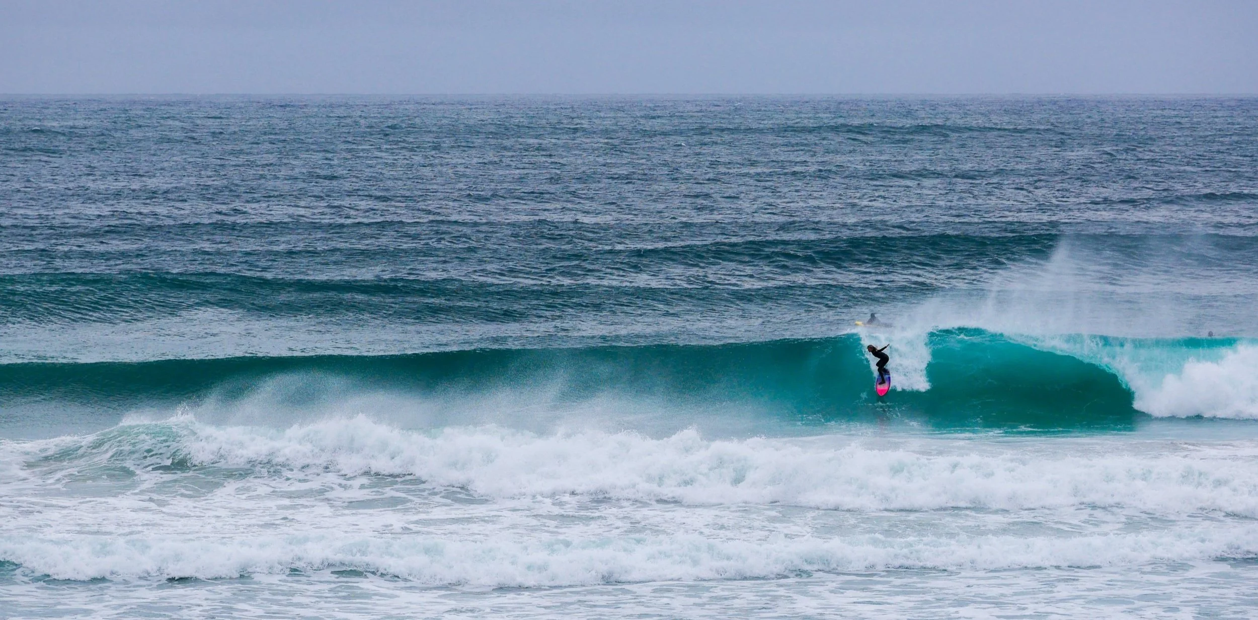 Surfer riding a wave in the ocean with a pink and blue surfboard.